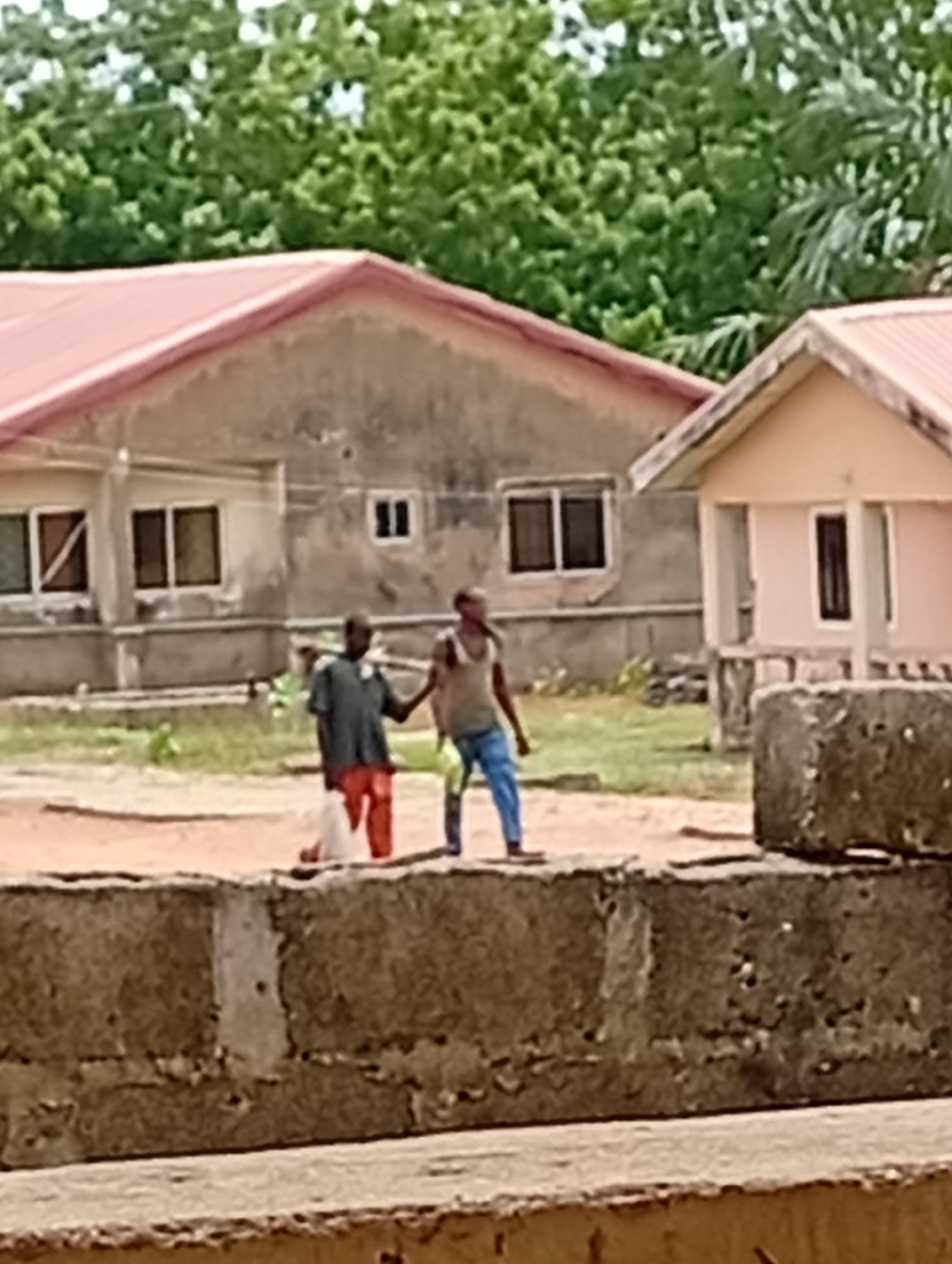 The image depicts two people walking in what appears to be Adankolo, Nigeria. One person is wearing a dark shirt and red pants, holding a white bag. The other person is wearing a tank top and blue pants. They are walking in front of two buildings with red roofs. The foreground shows a concrete structure. Green trees are visible in the background.
