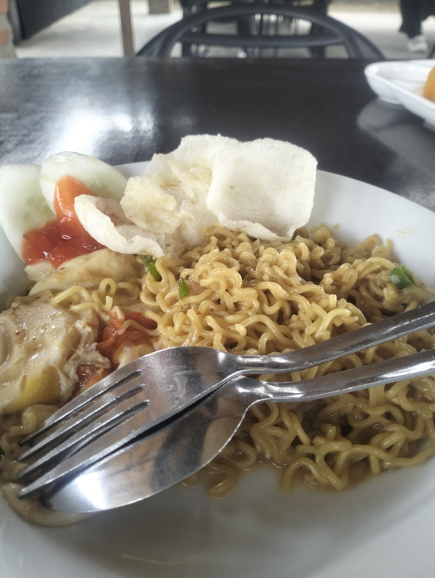 A close-up, eye-level shot shows a plate of Indonesian fried noodles served in a white bowl on a dark, reflective table. The noodles are curly and golden brown, mixed with a light-colored sauce. Partially visible are slices of cucumber with a dollop of red sauce, and several pieces of white crackers. A fork and spoon are resting on top of the noodles, angled from the left side of the frame. The background is blurred, showing dark furniture and a hint of an outdoor setting, suggesting the image was taken at a restaurant or cafe in Lhokseumawe, Indonesia.