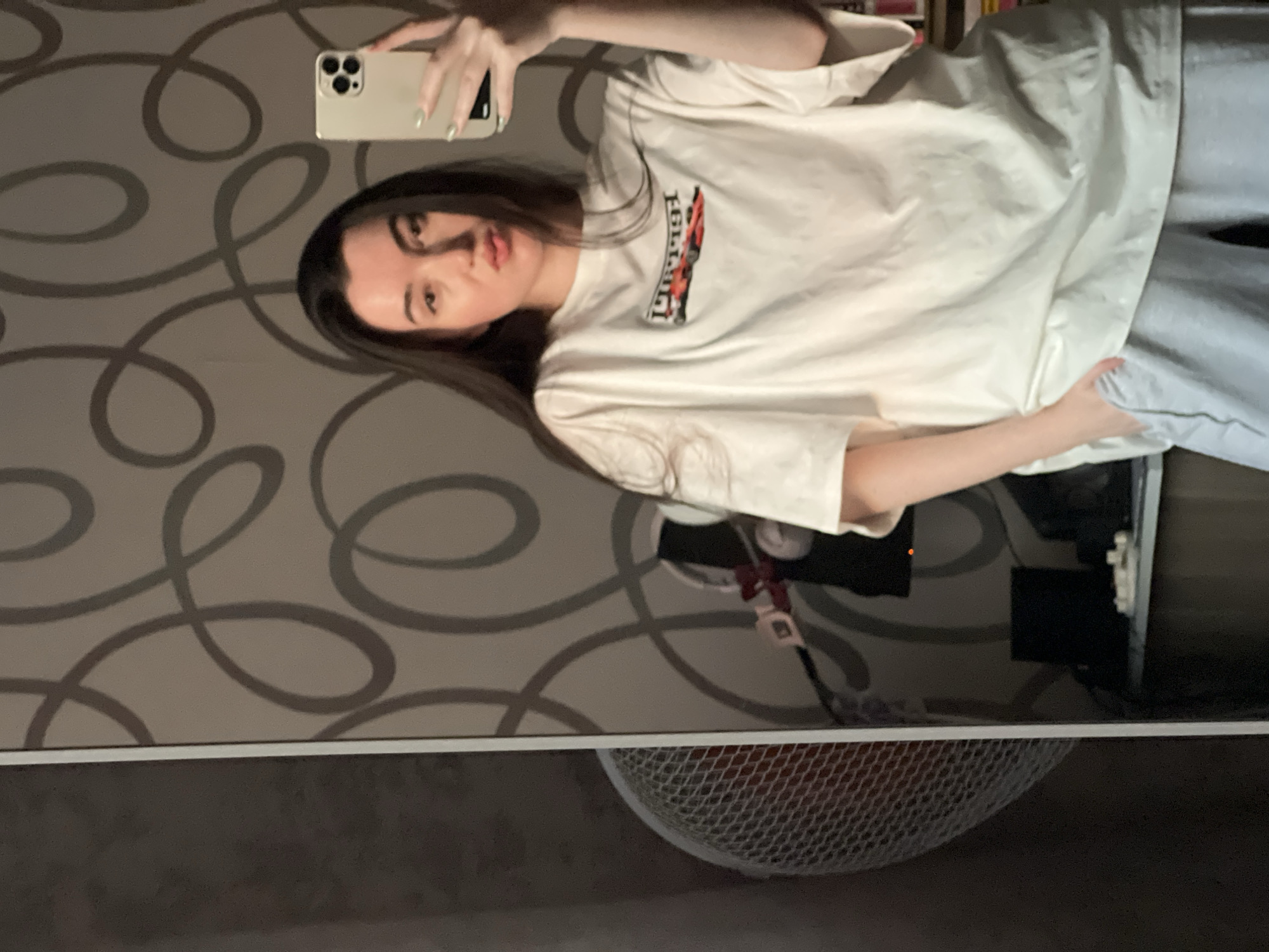 A young woman with long, dark hair is taking a selfie in a room. She is wearing a white t-shirt with a red car graphic and light gray sweatpants. Her right hand is holding a mobile phone and her left hand is in her pocket. The background features patterned wallpaper and a fan is visible on the left side of the image. The woman is positioned in front of a large mirror, reflecting her and part of the room. The room appears to be a domestic setting. There is no indication of the city of Kazan, Russia in the image.