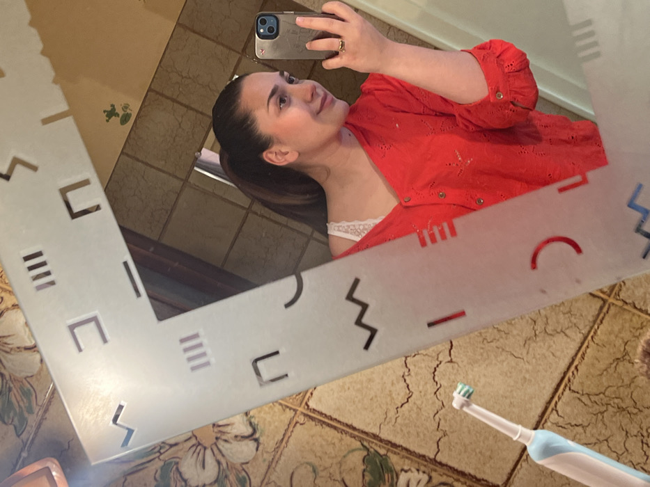 The image displays a mirror selfie taken within an interior space, likely a bathroom, located in Ancenis, France. The central subject is a female individual reflected in a rectangular mirror, which has a frosted border adorned with various geometric cut-out shapes including lines, squares, zigzags, and arcs. The individual possesses dark hair pulled back into a ponytail. She is wearing a red, long-sleeved, collared shirt with embroidered or eyelet details, unbuttoned to partially reveal a white lace undergarment. Her left hand holds a dark-colored smartphone, likely an iPhone due to the triple camera module visible, which is being used to capture the image. A gold-colored ring is present on her left ring finger. Her head is slightly turned to her left, and she exhibits a subtle smile. In the lower-left corner of the foreground, outside the mirror's reflection, a light blue and white electric toothbrush with a white brush head rests on a beige, patterned tiled surface. The visible background, both within and outside the mirror's reflection, consists of light-colored, possibly ceramic, tiles featuring a distressed texture and some floral or organic patterns.