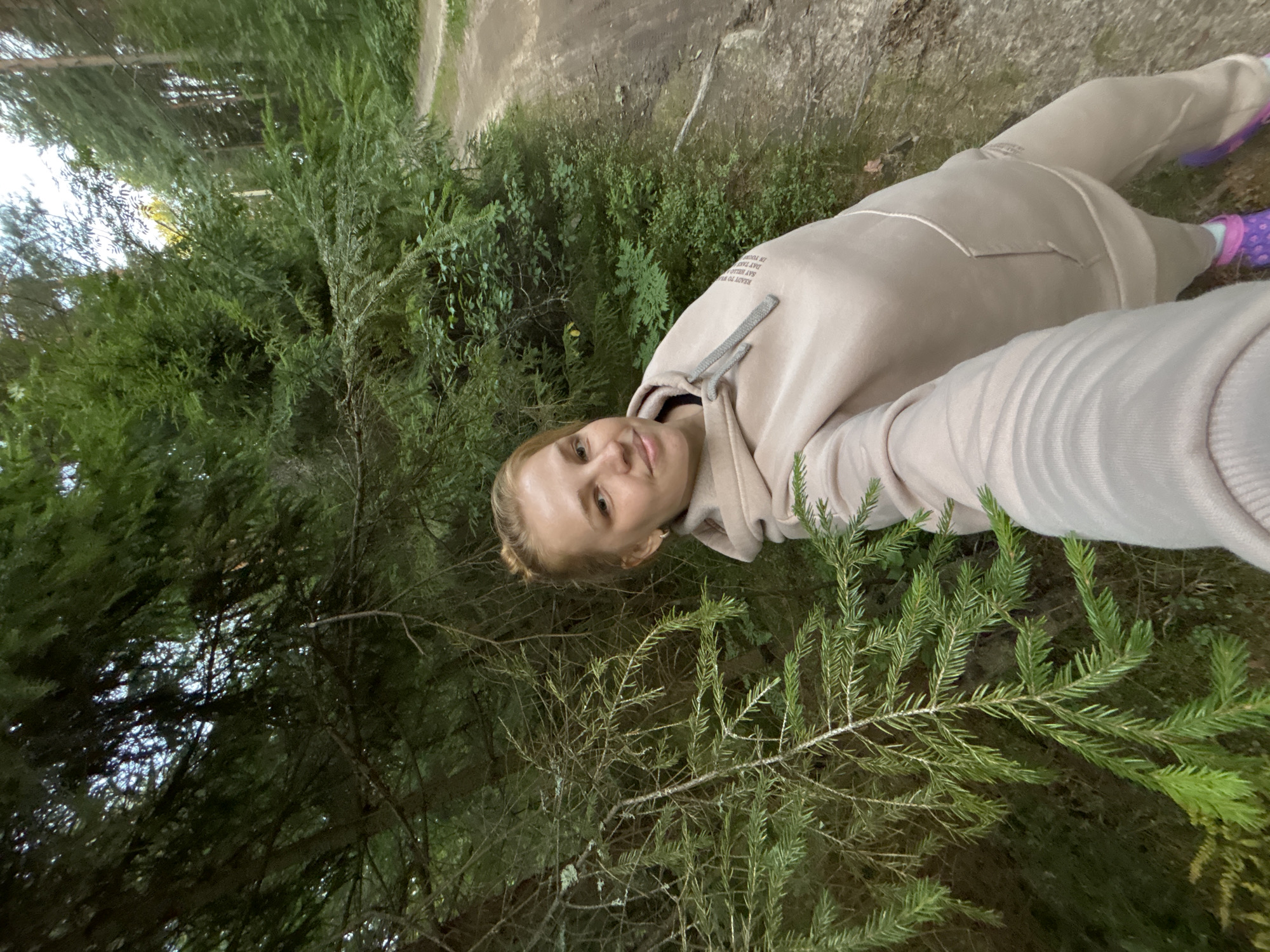 A woman is taking a selfie in a forest, with a dirt path to her right and dense evergreen trees surrounding her. She is wearing a light beige hooded sweatshirt and matching pants, with purple Crocs on her feet. Her hair is pulled back in a bun, and she is smiling at the camera. The trees are tall and thick, with needles covering the branches. The ground is covered with fallen leaves and pine needles. The sky is visible through the gaps in the trees, and it appears to be overcast.