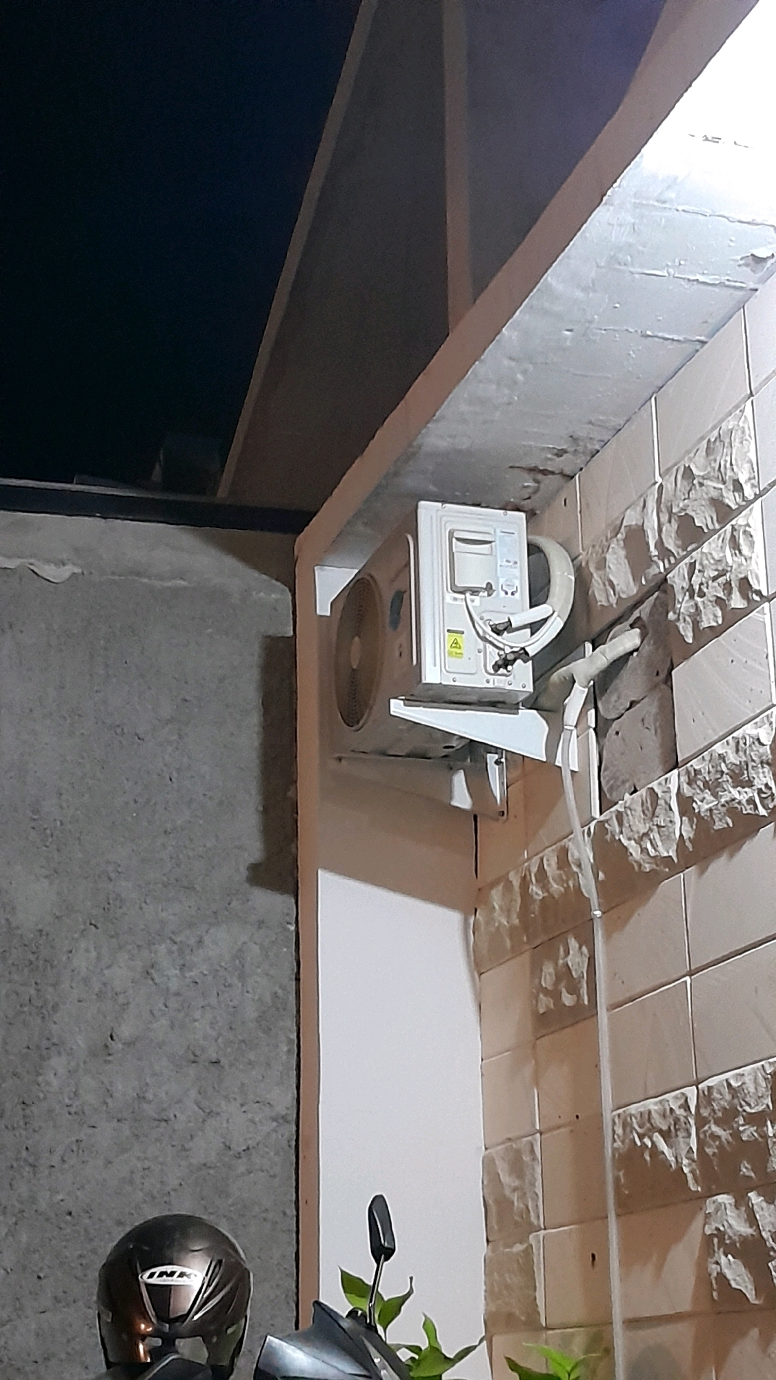 The image shows an outdoor air conditioning unit mounted on a textured wall, likely made of stone or brick. The unit is white and has a circular vent on its left side. Two pipes are connected to the unit, one insulated and the other appears to be a drainage pipe. The wall is light-colored with a rough, uneven texture. Above the wall, there is a dark overhang. In the lower left of the frame, a motorcycle helmet is visible, with part of a motorcycle and some green foliage. The scene appears to be at night or in low light conditions, as indicated by the dark background and the lighting on the air conditioning unit and helmet. The location is Pilangrejo, Indonesia.