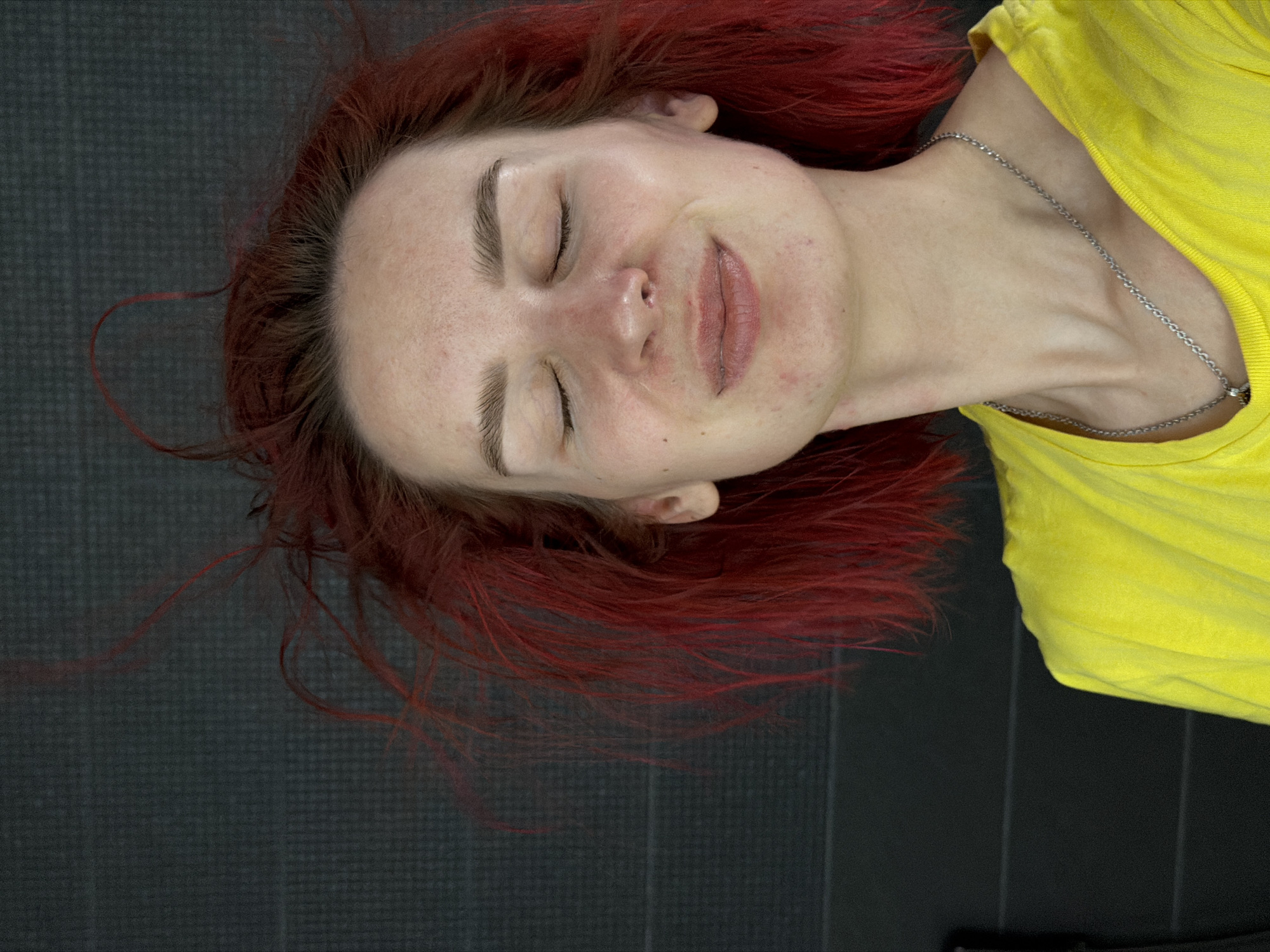 An overhead shot depicts a person, likely female, lying down with their eyes closed, suggesting rest or sleep. Her head is positioned towards the left of the frame, and her body extends to the right. She has distinctive red-auburn hair that spreads around her head on the dark surface. Her face shows a relaxed expression, with a gentle, closed-mouth smile. Visible facial features include light skin, well-defined eyebrows, and slightly parted lips. Her skin appears natural, with some minor freckles or imperfections.
She is wearing a bright yellow t-shirt, which is visible on the right side of the image, and a thin silver chain necklace around her neck. The background consists of a dark, textured surface, possibly a mat, suggesting an indoor setting. The overall mood conveyed is one of peace and contentment. No text is visible in the image. The location context for this media file is Chaikovsky, Russia.