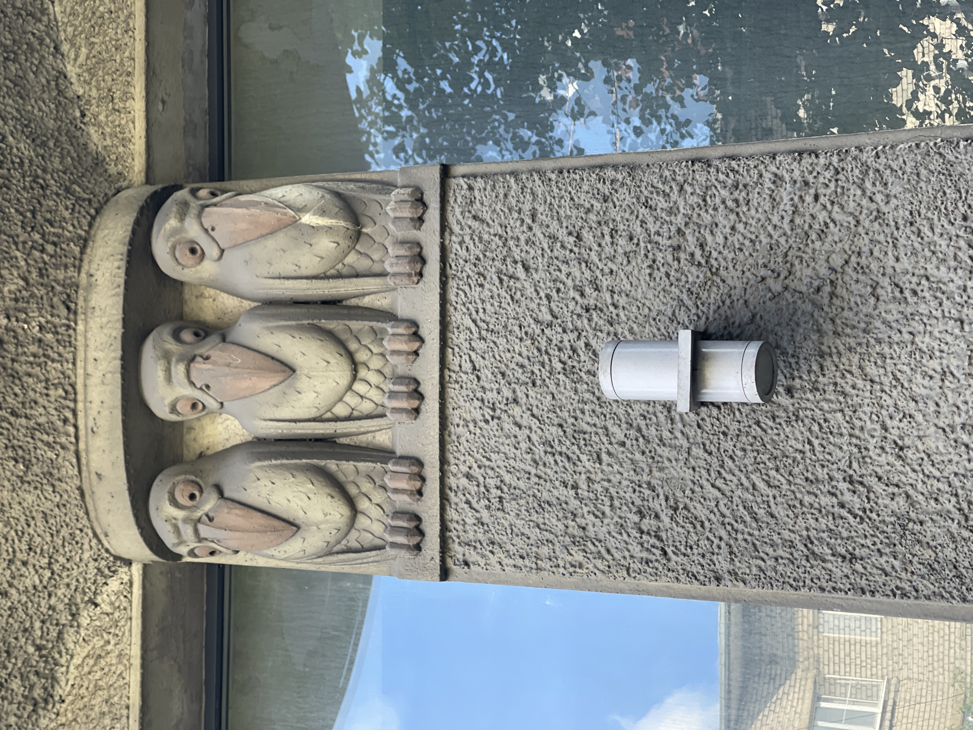 The image depicts a close-up of an architectural detail on the exterior of a building in Vyborg, Russia. The focal point is a decorative element featuring three carved bird-like figures in a row, mounted on a textured, grey stone column. Each figure has a prominent beak, round eyes, and appears to be standing on four small feet. The birds are positioned beneath an arch-like structure, also made of grey stone, which is textured with a rough, stucco-like finish. Below the bird figures, the column continues downwards, exhibiting the same rough grey texture. Attached to the column at mid-height is a cylindrical, silver-colored fixture, possibly a light or a conduit, with a protruding collar.
To the left of the column, a large window reflects a bright blue sky with some clouds. Further to the left, and also reflected in the window, is a portion of another building with light-colored brickwork and windows. To the right of the column, another window or glass surface reflects the dappled shadows and leaves of trees, suggesting an outdoor environment. The overall impression is of an architectural ornament integrated into the facade of a building in an urban setting.