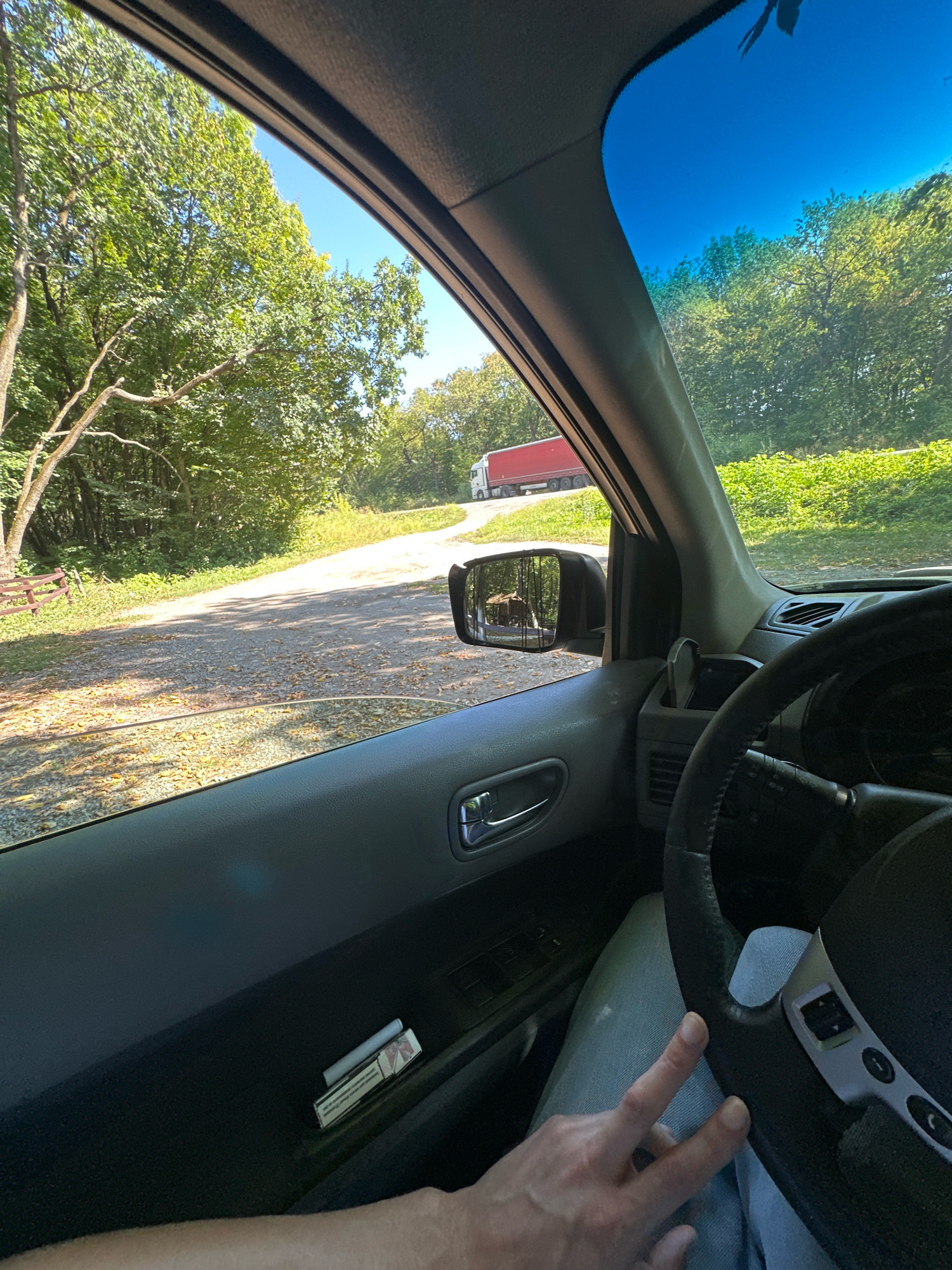 The image is taken from inside a vehicle, looking out through the windshield and side window. The exterior shows a rural, tree-lined road with a gravel surface. Sunlight filters through the leaves of mature trees on either side of the road. In the distance, a large red semi-trailer truck is visible on the road. A side-view mirror reflects a wooden structure or fence and trees. Inside the vehicle, the dashboard and steering wheel are visible, along with a hand resting on the steering wheel, suggesting the vehicle is in motion or parked. A pack of cigarettes and a lighter are placed in the door panel. The sky is clear and blue. The image does not provide any specific information that would allow for its location to be pinpointed to the city of Shatava, Ukraine.