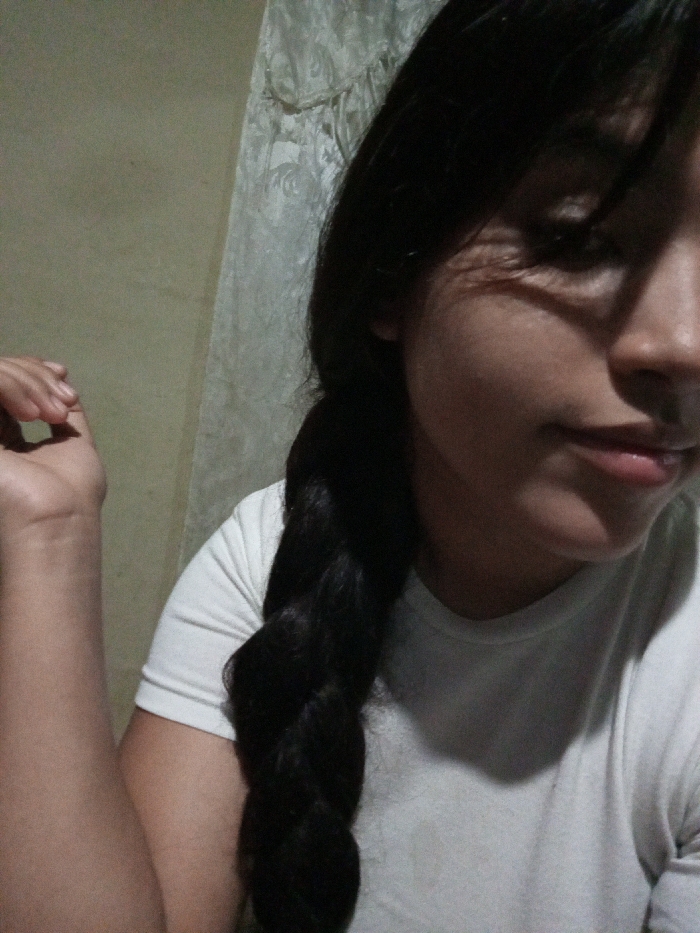 A young woman with dark hair braided down her chest wears a plain white t-shirt. Her eyes are cast down, and she has a soft smile. She holds her hand up in the air, bent at the wrist. A textured wall behind her gives way to a smooth pale surface in the background. Shadow lines her face and arms.