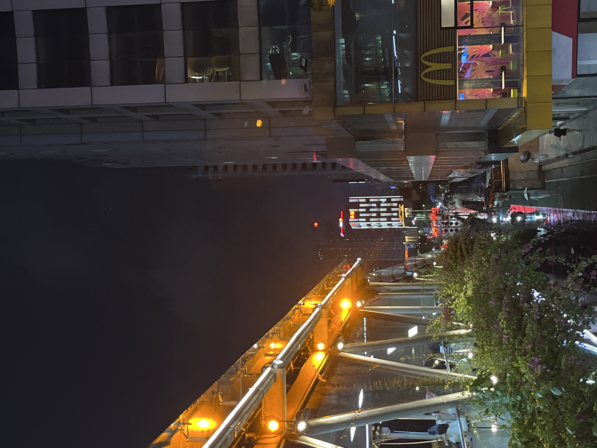 This is a nighttime photograph of an urban scene in Nanhai District, China. In the foreground to the left, a brightly lit elevated walkway or bridge is visible, with orange lights and railings. Lush green foliage with some pink flowers cascades down from the walkway. To the right, a modern building with a glass facade is prominent, featuring the golden arches of a McDonald's restaurant on a yellow facade on its lower level. Several tables and chairs are visible inside the McDonald's. A street runs below the walkway, with a few people walking, some holding umbrellas, suggesting it may be raining or has recently rained, as the ground appears wet. Other buildings and city lights are visible in the background, indicating a busy urban environment. The sky is dark and cloudy.