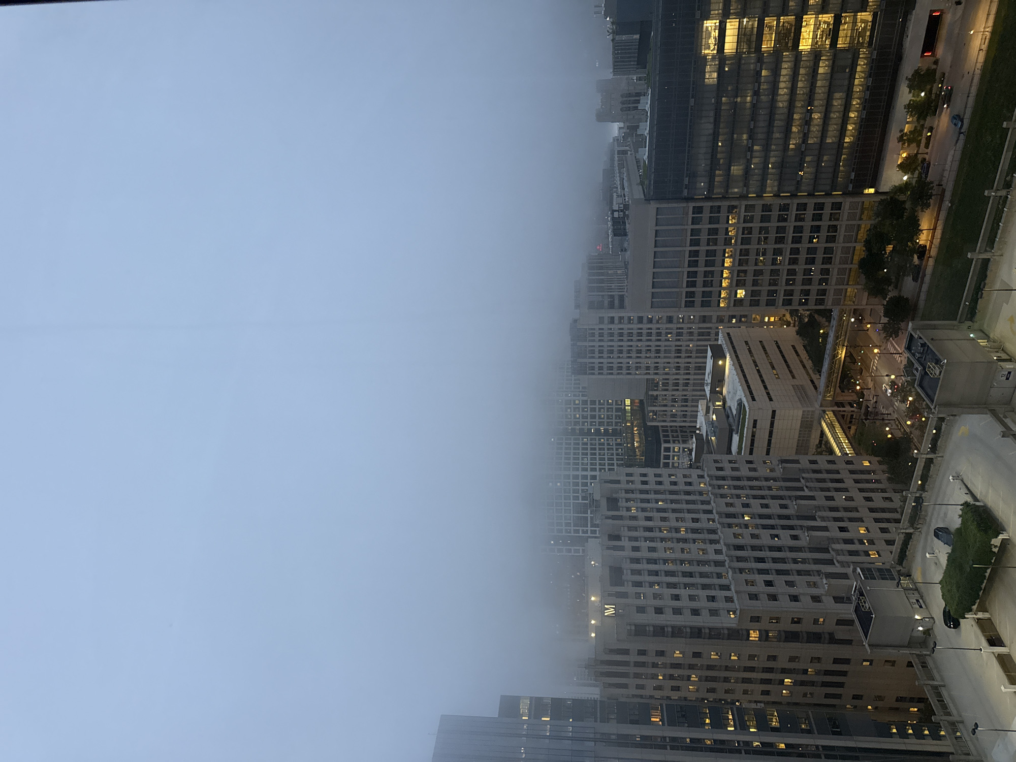 The image shows a cityscape in Chicago, United States of America, under a thick layer of fog. The view is from a high vantage point, looking down on several tall buildings of varying architectural styles. The sky is a uniform, pale blue-gray, obscured by fog which also blankets the upper portions of many buildings, obscuring distant structures. The foreground reveals a multi-level parking structure with parked cars and some greenery. Below the parking structure, street-level views show roads with vehicles, streetlights, and trees. The buildings themselves exhibit illuminated windows, suggesting occupancy within the structures. One building prominently displays a large, illuminated "M" logo. The overall atmosphere is muted and somewhat somber due to the fog and low light conditions, likely indicating early morning or late evening.