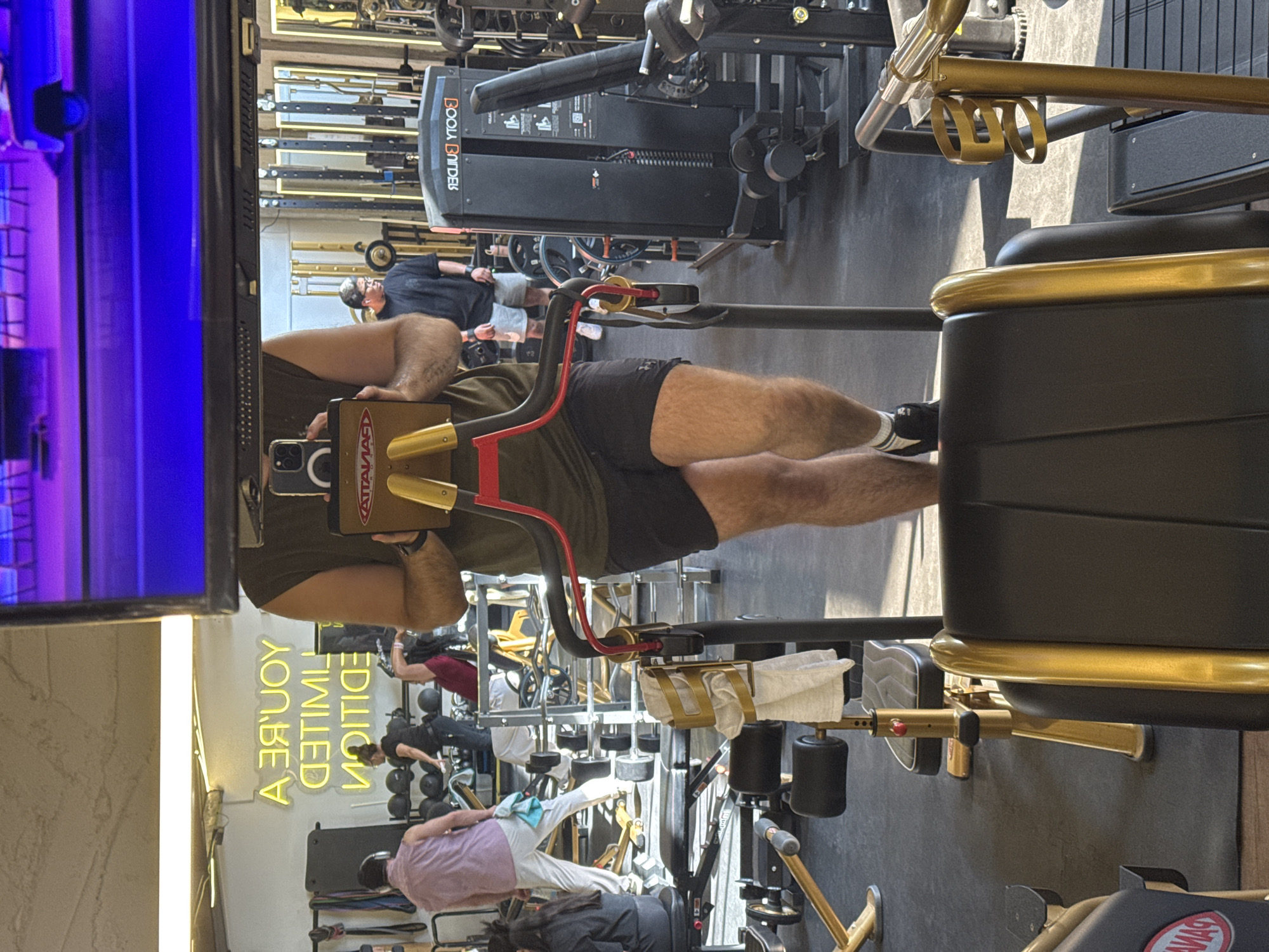 The image captures a person in a gym, likely taking a selfie with a leg press machine in the foreground. The machine has a black seat with gold accents. Other gym equipment, including a cable machine by Body Builder, is visible in the background, along with multiple other gym-goers. The gym appears to have a neon sign on a wall reflecting in a television screen. Puebla, Mexico is not mentioned.