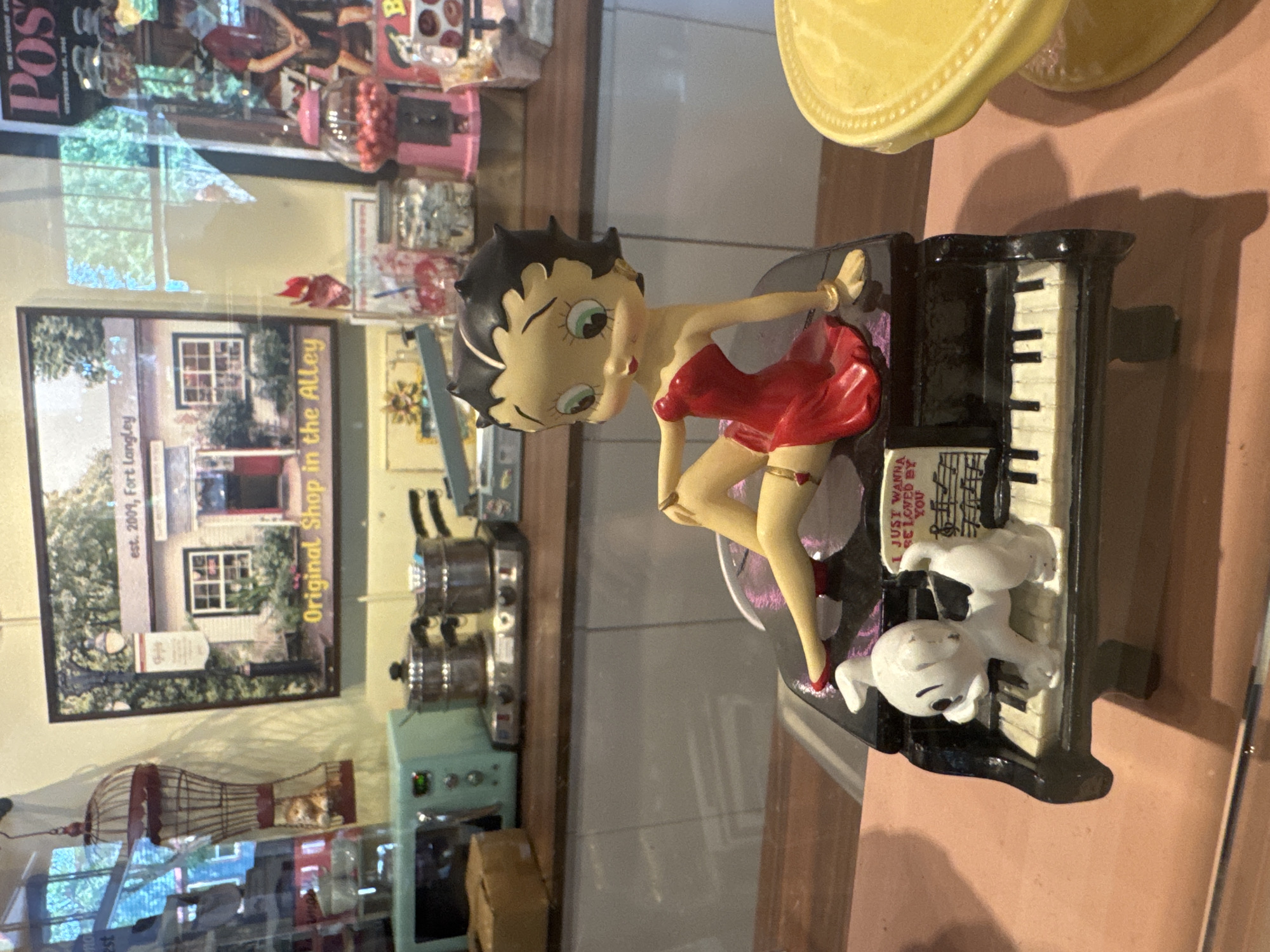 The image depicts a retail display, likely inside a shop. A Betty Boop figurine, dressed in a red dress, is posed atop a black miniature piano. A small white dog figurine stands on the piano's keys. A sheet of music is visible on the piano. The background shows various items, including a framed picture of a building with text indicating "est. 2009, Fort Langley" and "Original Shop in the Alley." There are also gumball machines, glassware, and kitchen appliances visible. The overall impression is of a quirky, nostalgic retail environment, consistent with a gift shop or novelty store. The mention of "Fort Langley" places the location in the Township of Langley, British Columbia, Canada.