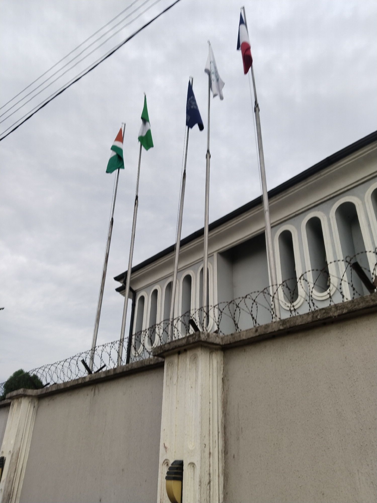 In Uyo, Nigeria, five flags fly on tall poles in front of a pale gray, modern building with arched windows. From left to right, the flags are a tricolor of orange, white, and green; a green and white flag; a blue and white flag; a white flag with blue markings; and the French tricolor flag. The building is bordered by a concrete wall topped with coiled barbed wire. Overhead, dark power lines traverse a cloudy sky. The building's facade is decorated with white trim around the arched windows. The wall in the foreground features decorative pilasters on its left and right sides, and two light fixtures are visible near the bottom corners of the image. The overall atmosphere suggests an official or diplomatic location.