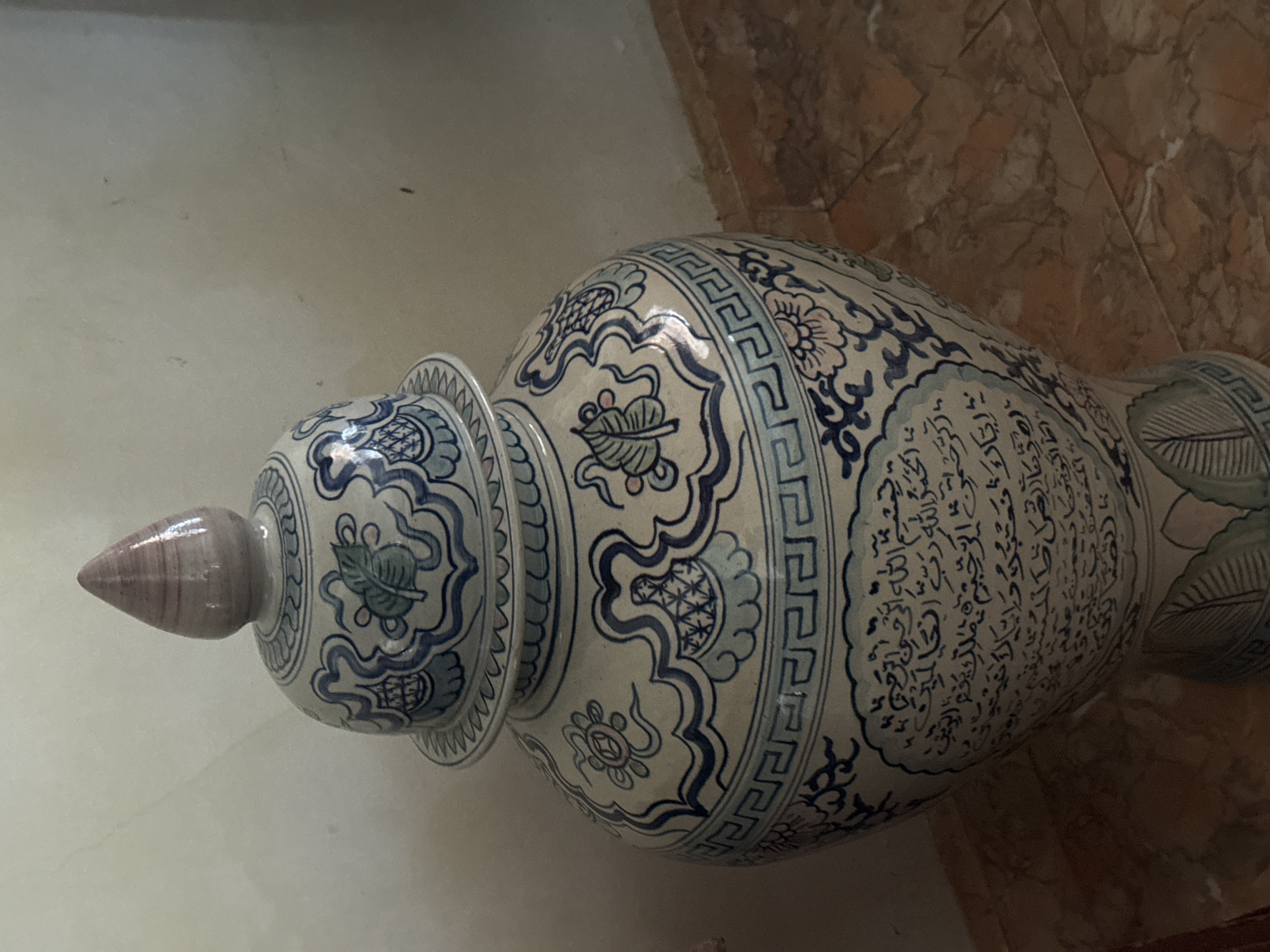 The image shows a large ceramic jar with a lid, decorated with blue and white patterns. The jar features floral motifs, geometric borders, and Arabic calligraphy. It is placed on a tiled floor, possibly in a domestic setting. The background is a plain, light-colored wall. There is no visual information in the image that can identify the specific location as Bireuenmeunasahblang, Indonesia.