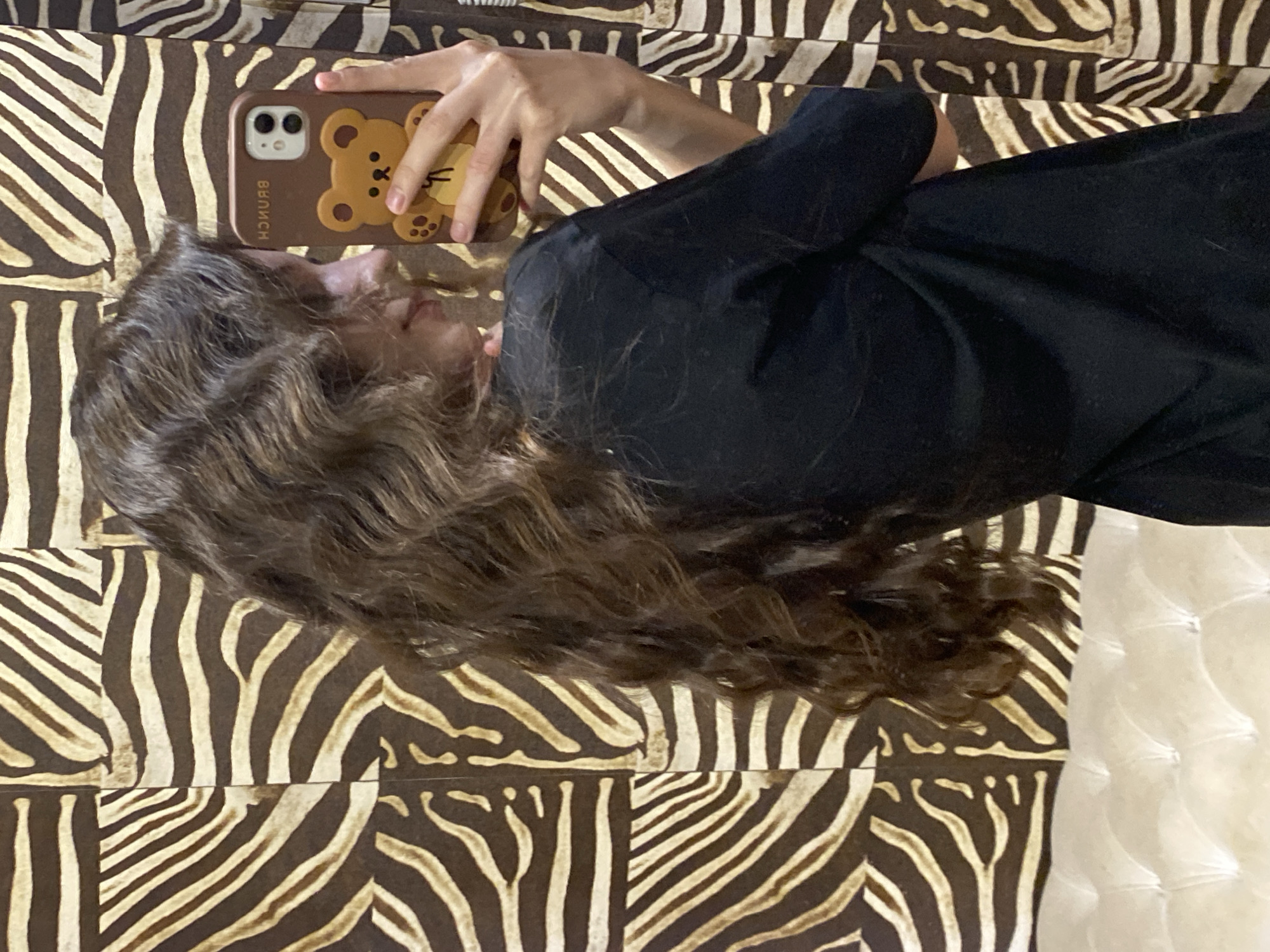 A woman with long, wavy brown hair is lying on a zebra-print surface, possibly a bed or sofa. She is wearing a black shirt. She is holding a brown iPhone with a teddy bear design on the case in her right hand. The image does not contain information about Votkinsk, Russia.
