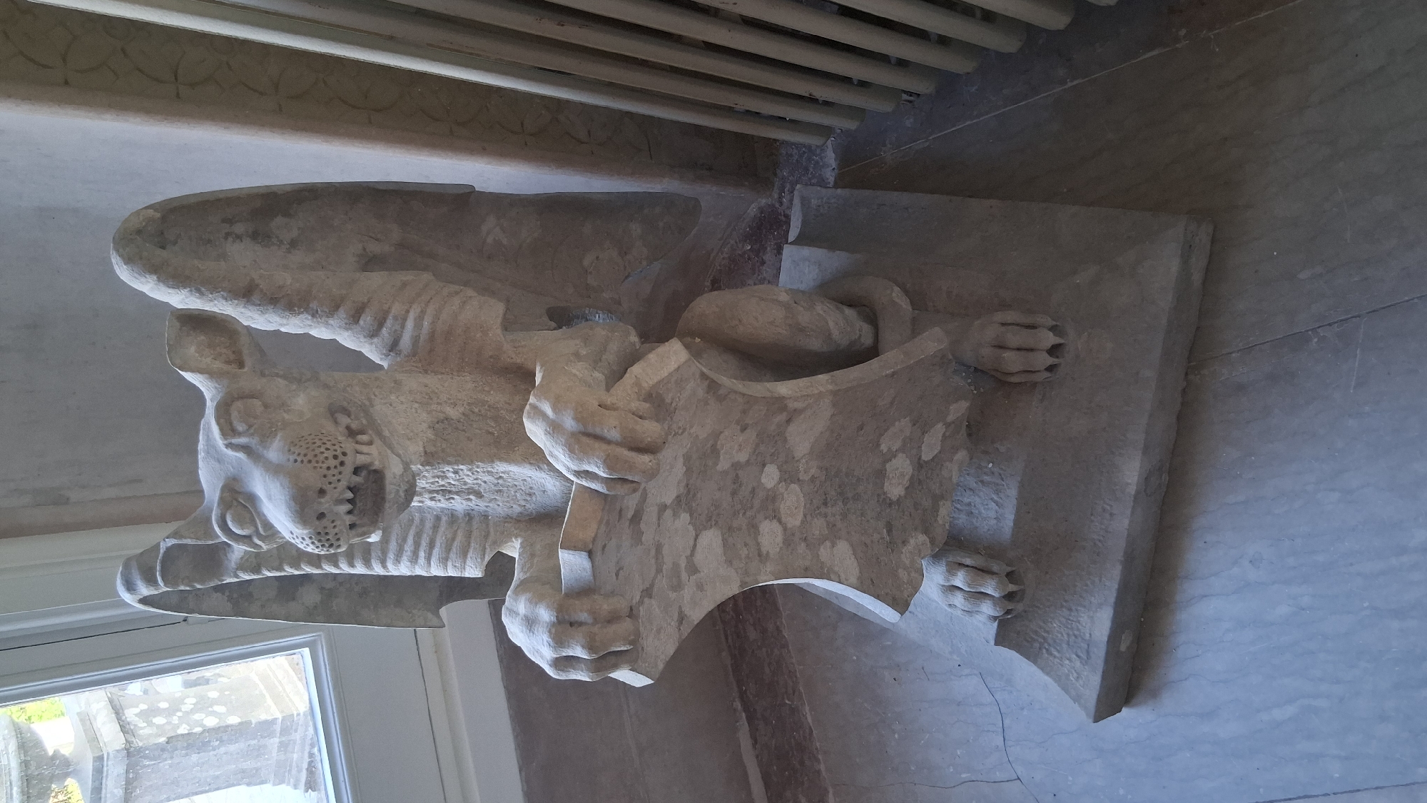 A gargoyle statue carved from light-colored stone is positioned on a stone floor, facing forward. The gargoyle has wings, a dog-like head with bared teeth, and clawed paws holding a shield-like object. The statue is situated indoors, with a window and part of a building facade visible to the left, and a radiator or similar structure to the right. The flooring appears to be made of large stone tiles. The background wall is textured and light-colored. The location is identified as Sintra, Portugal, specifically within the parish of Santa Maria e São Miguel, São Martinho e São Pedro de Penaferrim.
