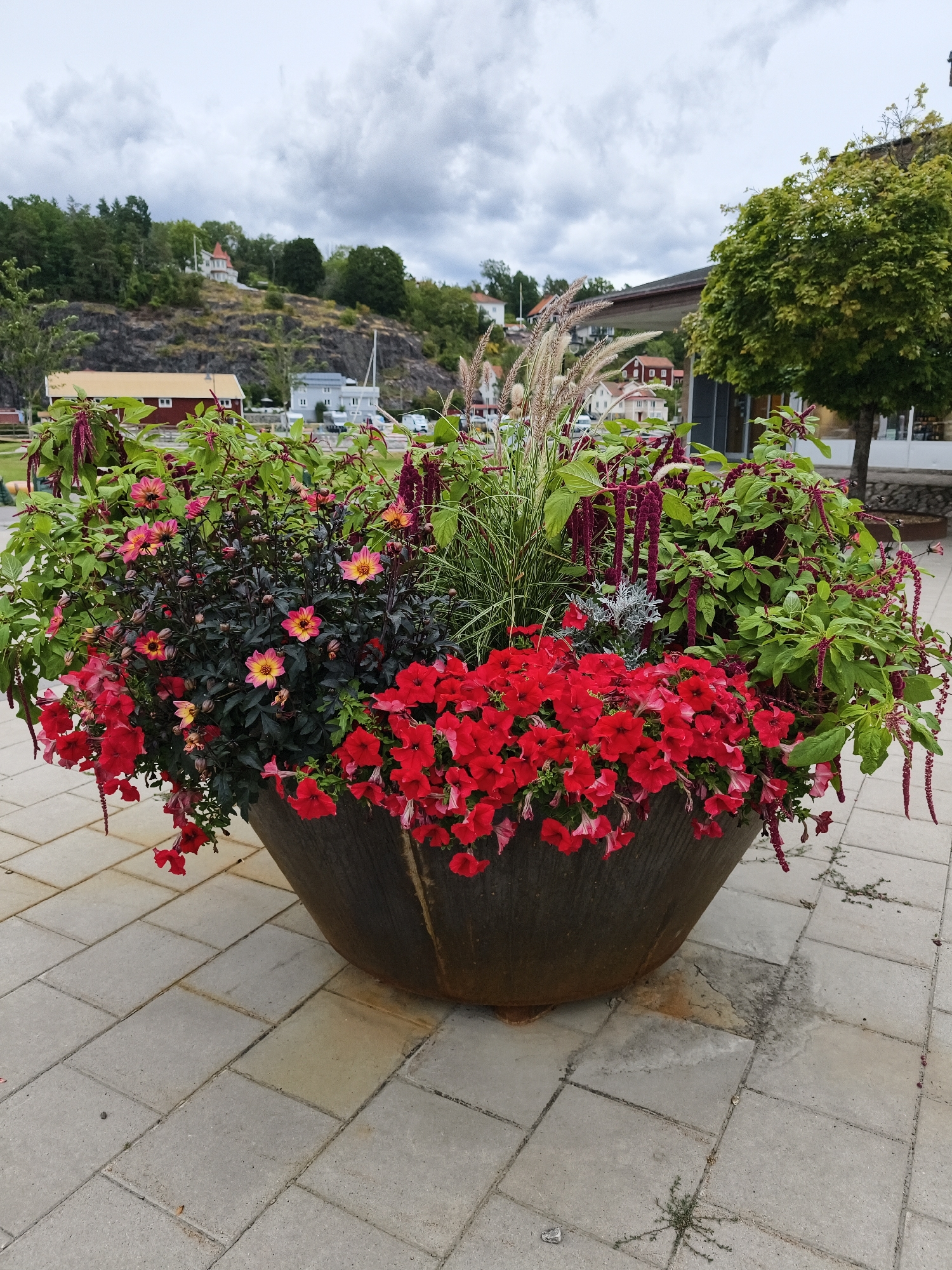 A large, weathered metal planter overflows with a lush arrangement of red petunias, pink and yellow dahlias, and trailing red amaranth. The planter sits on a light gray paver stone patio. In the background, a green tree stands to the right of the planter, and a cluster of buildings is visible on a hillside. The sky is overcast with heavy clouds. This scene appears to be located in the town of Valdemarsvik, Sweden, possibly near a waterfront area, given the presence of what looks like a sailboat in the distance. There are no people visible in the image.