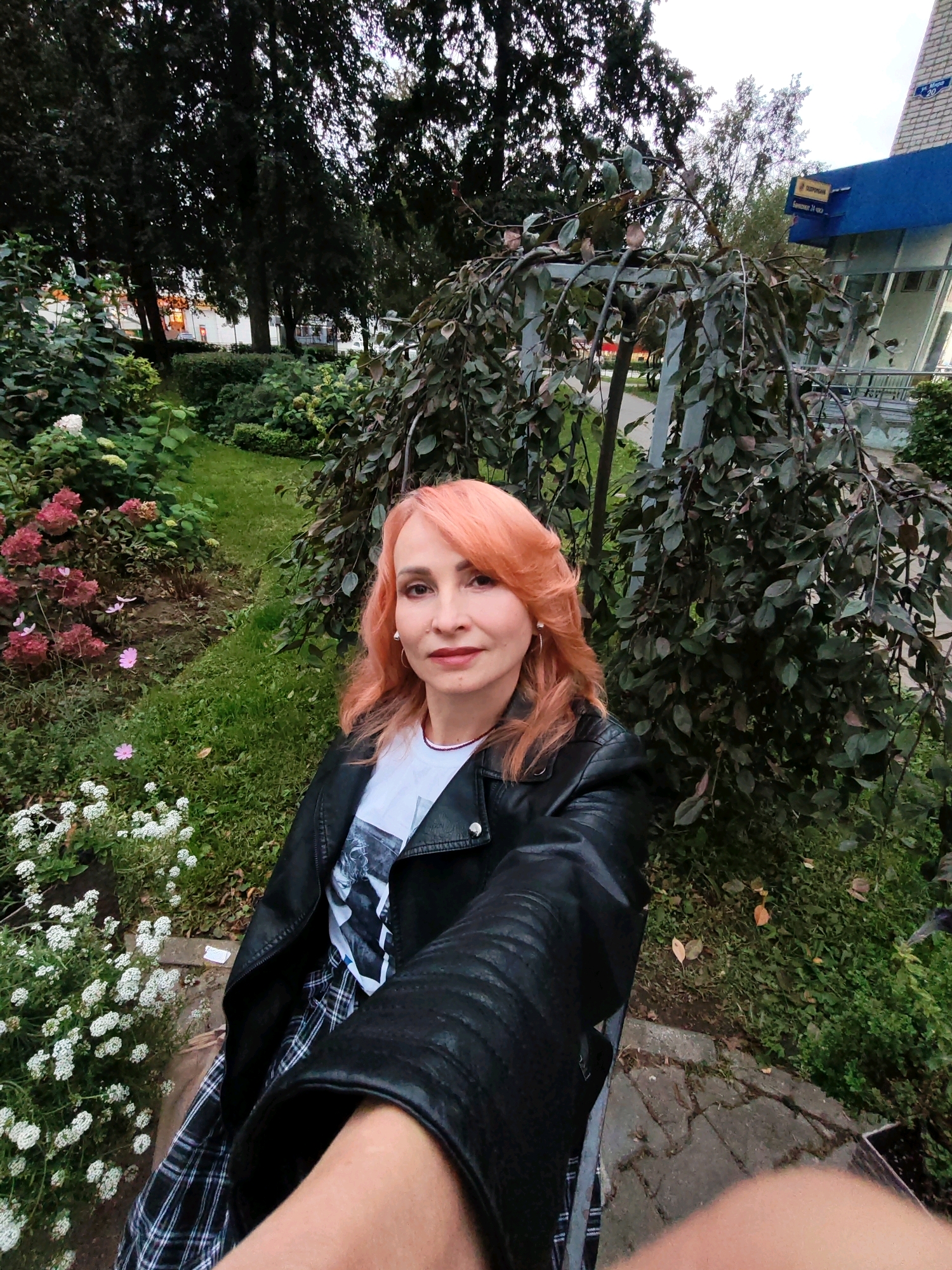 The image is a selfie taken outdoors in a garden with a building in the background. A woman with salmon-colored hair is the primary subject. She is wearing a black leather jacket over a white t-shirt and a plaid skirt. She is seated, with her right arm extended towards the camera, holding the phone. The background features a variety of plants and flowers, including pink hydrangeas, white alyssum, and a weeping tree with dark leaves. A paved walkway is visible in the foreground. In the background, a blue and white building with signage is partially visible. Based on the signage, the location is identified as Chaikovsky, Russia, specifically near a bank or financial institution. The overall lighting suggests it is daytime, possibly overcast.