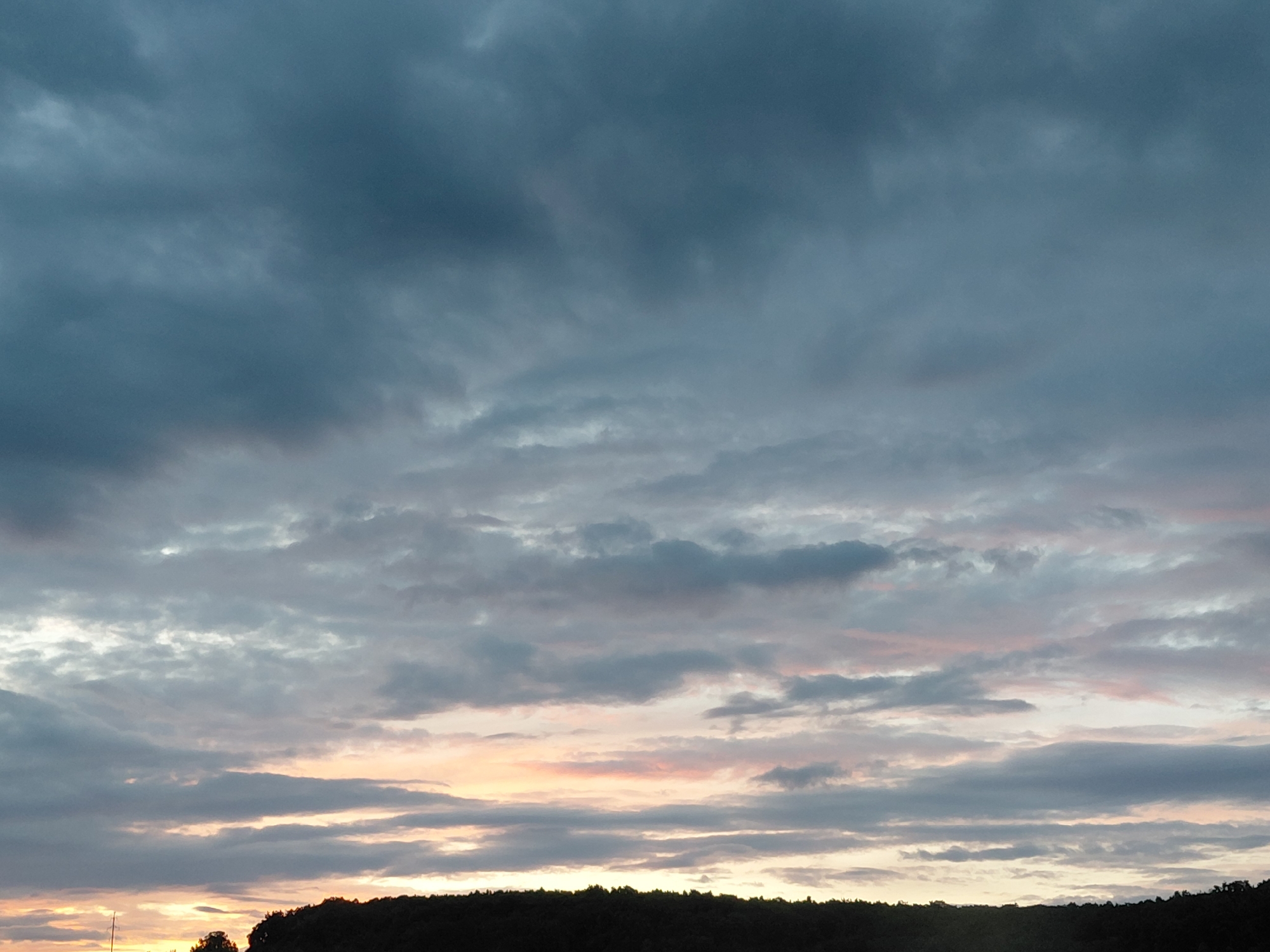 The image depicts a wide, overcast sky with a dark, silhouetted tree line in the foreground, likely a wooded area. The clouds are dark gray and heavy, with streaks of lighter gray and hints of pink and orange from a setting or rising sun peeking through. The lighting suggests either dusk or dawn. The terrain is uneven, with the dark mass of the trees obscuring any finer details of the ground. The scene appears to be in a natural or semi-natural setting, possibly a park or forest on the outskirts of Novokuybyshevsk, Russia. There are no people or specific identifiable structures visible in the frame.