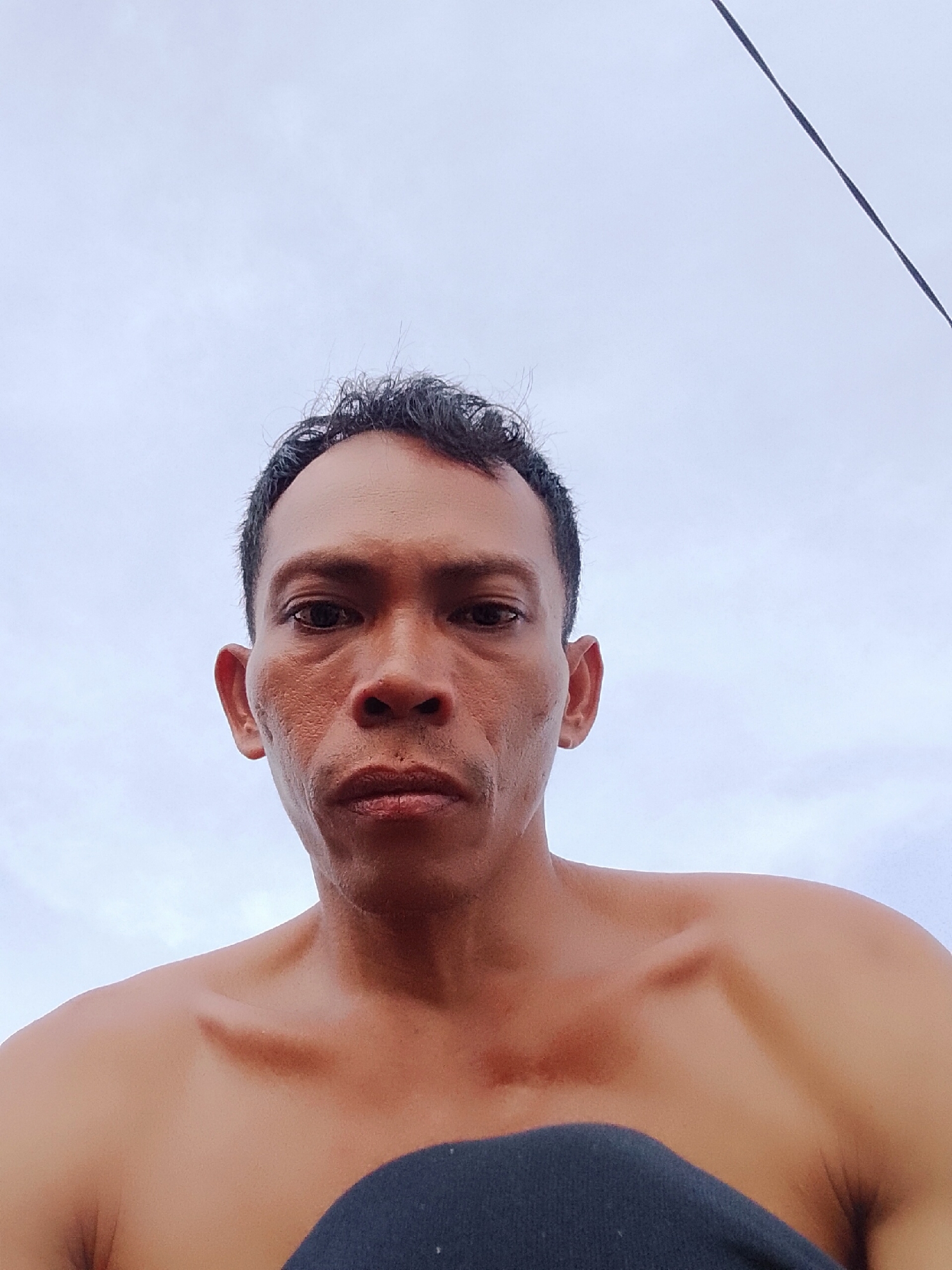 A medium-shot, eye-level photograph depicts a shirtless man with short, dark hair. His skin appears damp. He is centered in the frame and faces the camera directly. Above him is a cloudy sky and a powerline that runs diagonally. Based on the prompt, the location is Bukit Ketok, Indonesia. No other objects or subjects are clearly visible.