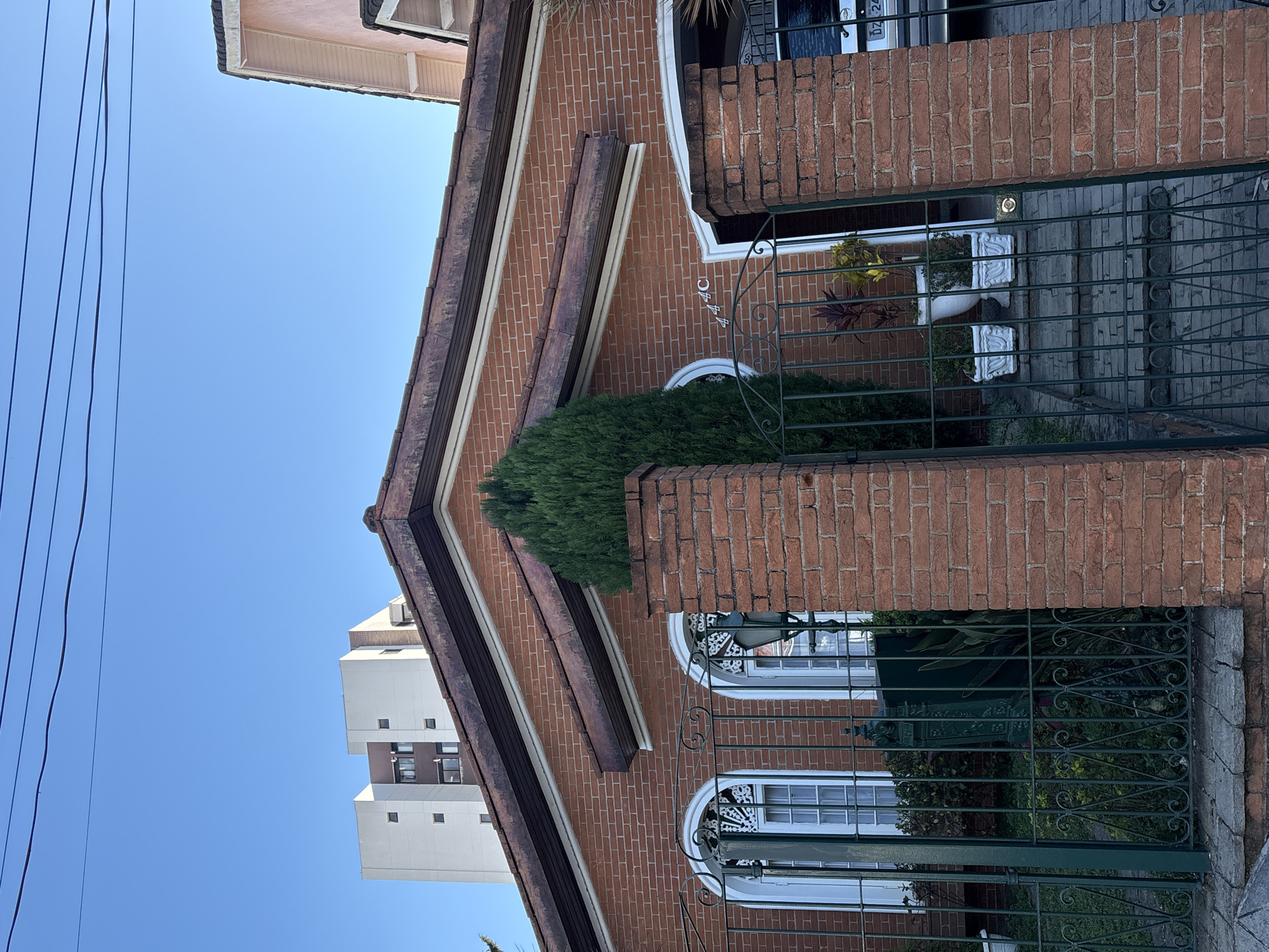 This is an eye-level, exterior shot of a brick house in São Paulo, Brazil, featuring a clear blue sky and overhead power lines. The house has a prominent red brick facade with arched white-framed windows and decorative iron gates. A tall, green evergreen tree stands centrally in front of the house, obscuring part of the facade and a circular decorative window. To the left of the house, a modern, multi-story building is visible. The entrance to the house is visible through an open gate, revealing a few potted plants on a set of brick stairs. A partially visible car with a license plate that reads "DZ 24" is parked to the right of the entrance. The overall scene depicts a residential street on a sunny day.