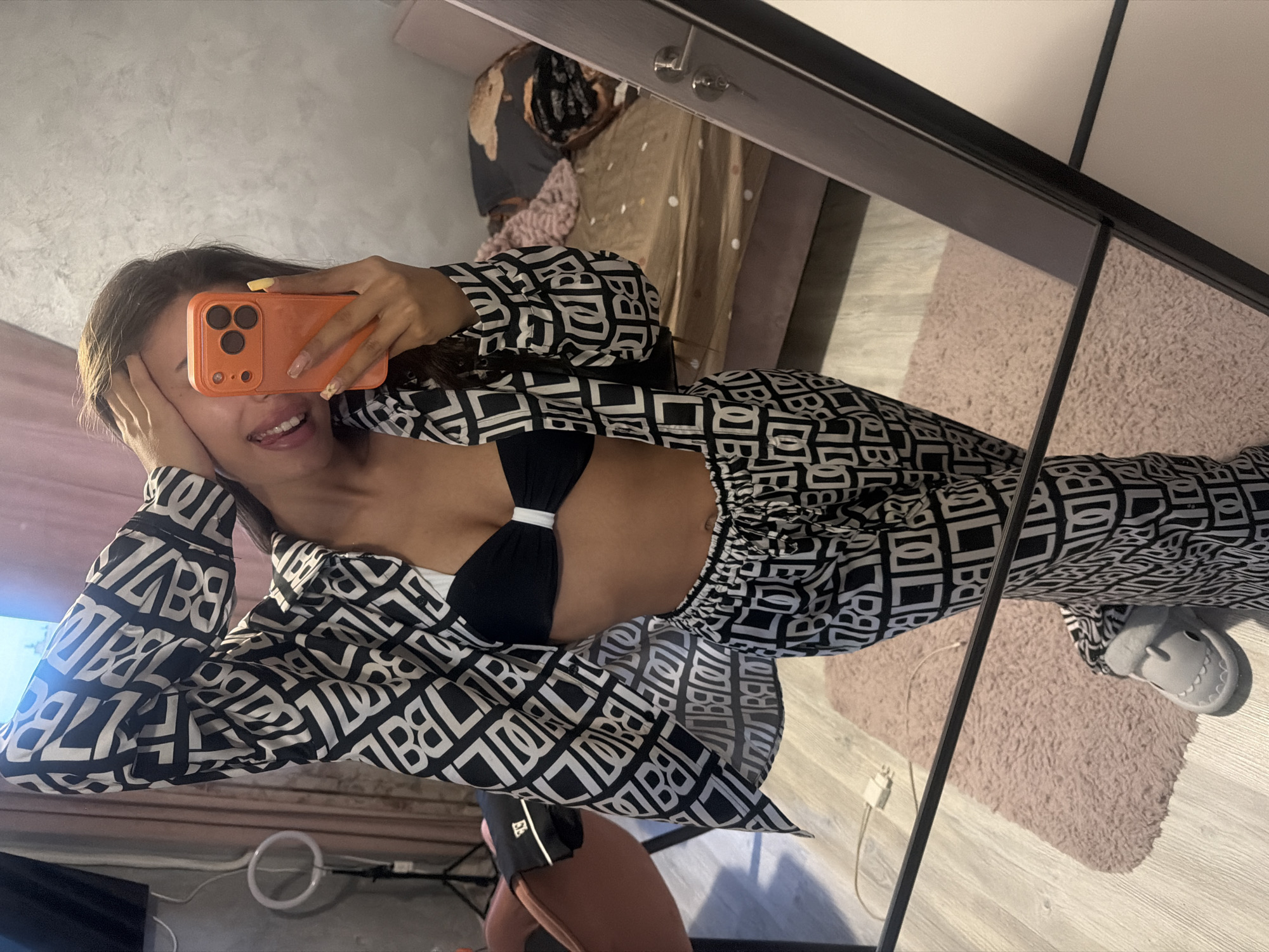 A young woman with long brown hair is taking a selfie in a mirror. She is wearing a black bikini top and a matching black and white patterned pajama set. The pajama set includes a loose-fitting button-down shirt and wide-leg pants. The woman is holding her phone, which has an orange case, in her right hand, and her left hand is resting on her head. She is smiling and sticking her tongue out. The background shows a bedroom with a bed, a pink rug, and a ring light on a stand. The image was taken indoors, likely in a residential setting, and there is no indication of its location in Burgas, Bulgaria.