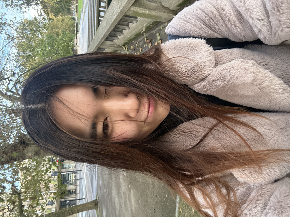 A young East Asian female is depicted in a close-up, chest-up selfie. Her long, dark brown hair, with lighter reddish-brown ends, is parted centrally and slightly wind-blown across her face. She smiles with closed lips, her right eye (viewer's left) partially closed in a wink, while her left eye is open. She wears a black high-collared garment beneath a light beige, fuzzy, textured coat. A black strap, likely from a backpack, is visible over her right shoulder.
The background reveals an urban environment. To the left, a paved street with a visible crosswalk and bare deciduous trees lines the sidewalk. Multi-story buildings with windows are visible further in the distance, and a red traffic light signal is partially obscured by a tree branch. To the right, a distinctive concrete balustrade with vertical slats separates the foreground from a green area featuring deciduous trees, some retaining green leaves, and scattered fallen leaves on the ground.
The scene is set outdoors during daylight. The architecture, particularly the concrete balustrade and urban planning, indicates the city center of Le Havre, France, likely in the vicinity of the Bassin du Commerce or the Hôtel de Ville, an area characterized by Auguste Perret's post-war reconstruction.