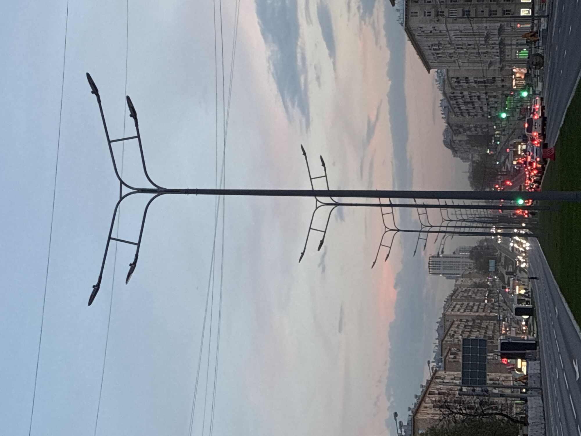 The image shows a street in Moscow, Russia, lined with tall buildings on both sides. A row of stylized streetlights with horizontal arms runs along the side of the road. Traffic is visible on the street, including cars and traffic lights. The sky is overcast with clouds.