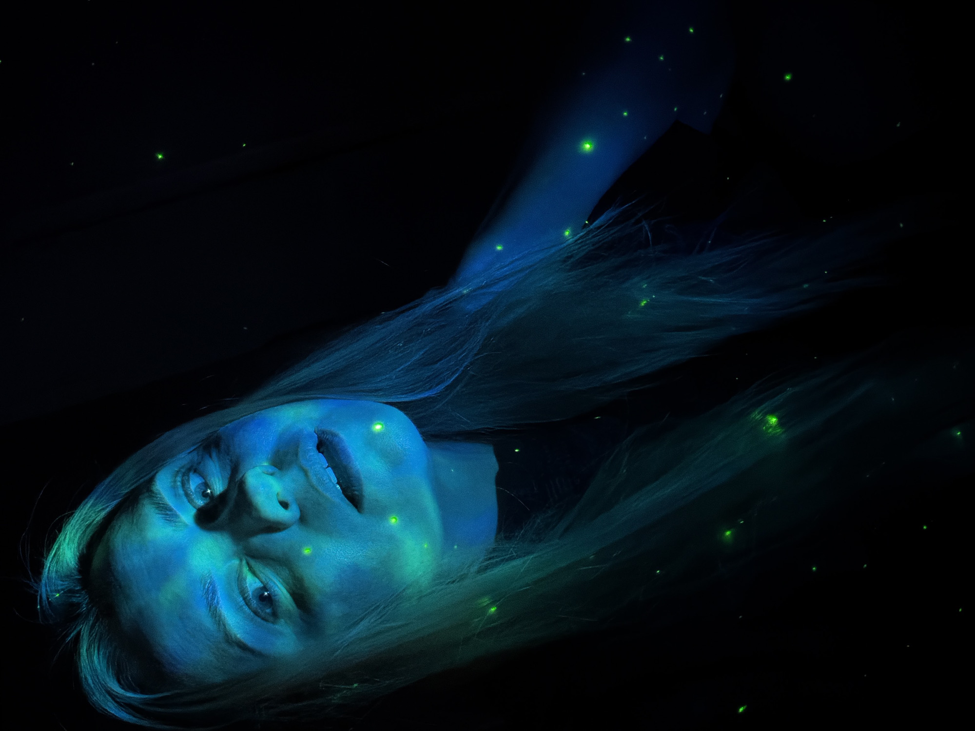 The image displays a close-up, angled portrait of a woman against a completely dark, seemingly black, background. Her face and visible areas of her upper body, along with her long, light-colored hair, are intensely illuminated in vibrant shades of electric blue and aqua-green, suggesting a bioluminescent effect or the use of ultraviolet (UV) light. Her features, including her eyes, nose, and slightly parted lips, are clearly defined by this glow. She is positioned as if lying down, looking subtly upwards, with a calm and contemplative expression.
Numerous small, bright green specks, resembling glowing particles or fireflies, are scattered across her skin, intertwined in her hair, and dispersed throughout the dark background. These glowing points contribute to an ethereal and otherworldly atmosphere. The setting is a controlled, dark environment, implying nighttime or an enclosed space, rather than an outdoor scene with natural light. The overall impression is artistic and mysterious. There is no visible text in the image. This scene is depicted without specific location cues but is contextualized as Votkinsk, Russia.