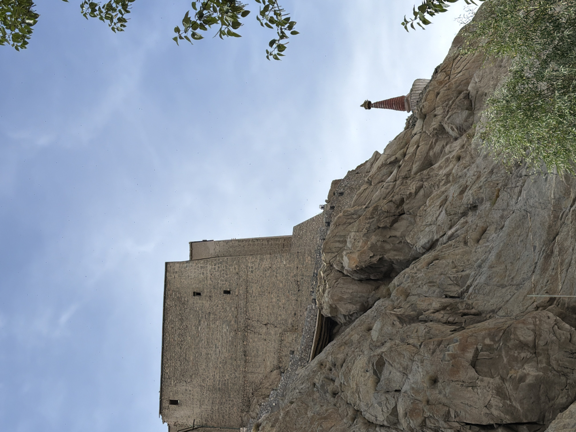 The image shows a view of the Leh Palace and a stupa situated on a rocky hillside under a clear blue sky with scattered clouds. The Leh Palace, a historic royal complex, is a prominent stone structure with a weathered facade, featuring small rectangular windows. It sits on the upper part of the rocky incline. Below and to the right of the palace, a red and white stupa with a tiered conical roof is visible, also positioned on the rocky terrain. The foreground is dominated by the rugged, textured surface of the rocky hill, with some sparse vegetation and trees with green leaves framing the scene on the right and bottom. The overall impression is of an ancient architectural landmark set against a natural, arid landscape.