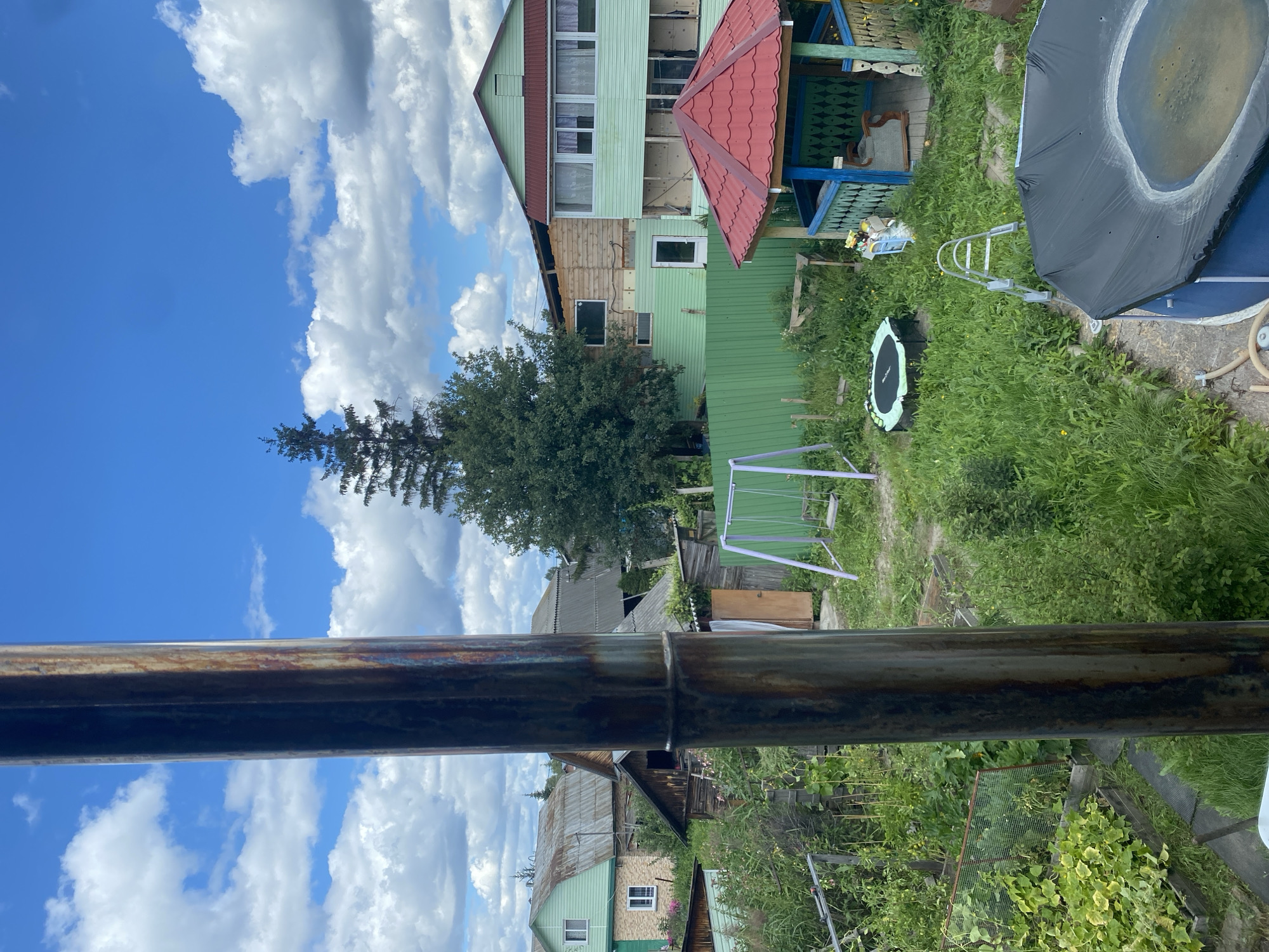 The image depicts a backyard scene in Krivodanovka, Russia, under a partly cloudy sky. In the foreground, a thick, rusty metal pipe runs vertically, obscuring a portion of the view. The backyard is filled with lush green grass and overgrown vegetation. Several objects are visible, including a small trampoline, a silver ladder leaning against the grass, a children's swing set with a purple frame, and a blue, round above-ground swimming pool covered with a black tarp. To the right, a hexagonal gazebo with a red roof and blue and green painted posts stands among the greenery. Behind the gazebo, a multi-story building with light green siding and a dark red roof is partially visible. Various other wooden structures and houses with pitched roofs are scattered throughout the background, suggesting a residential area. There are no people or animals visible in the image.