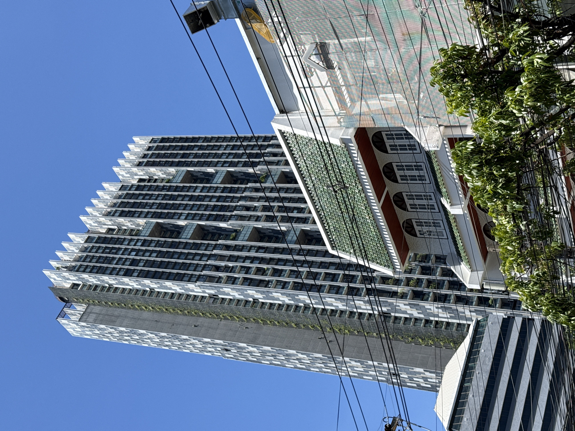A low-angle shot captures a portion of a modern skyscraper with a distinctive, stepped facade, a traditional building with a green tiled roof, and another contemporary building with horizontal striped windows, all set against a clear blue sky. The scene is in Bangkok, Thailand. Overhead, a dense network of power lines and utility cables crisscrosses the frame, obscuring parts of the buildings. Lush green foliage from trees in the foreground further obscures the lower sections of the buildings and power lines. The skyscraper features a patterned exterior with alternating light and dark vertical elements and recessed balconies. The traditional building exhibits arched windows with white frames and a dark trim under the eaves. The striped building has a sleek, dark exterior with evenly spaced horizontal white bands. The overall impression is a juxtaposition of modern and traditional architecture within a busy urban environment characterized by abundant overhead wiring. No people are visible.