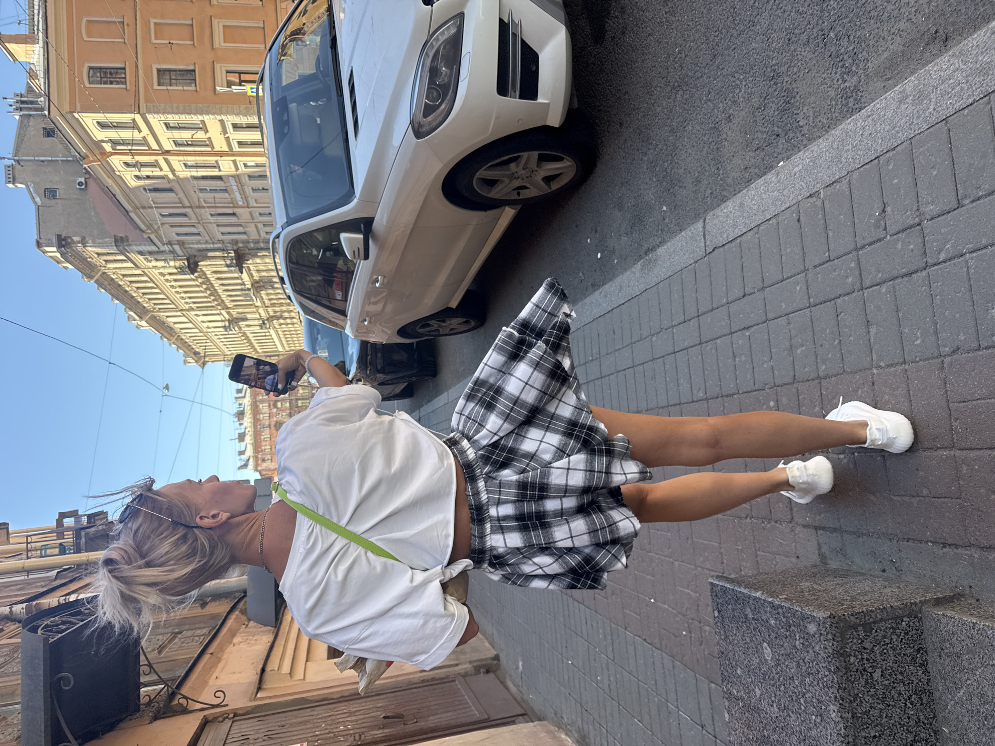 A woman with blonde hair pulled up in a bun stands on a cobblestone sidewalk, holding a smartphone to take a selfie. She is wearing a white t-shirt and a black and white plaid skirt. Her legs are bare, and she is wearing white sneakers. A lime green strap is visible across her chest, likely from a bag or purse.
Behind her, a white SUV is parked on the street. The buildings on the other side of the street are tall and appear to be old architecture, typical of Saint Petersburg. The sky is blue and clear, suggesting a sunny day. The scene appears to be in a city, likely during daylight hours. The woman's focus on her phone suggests she is documenting her experience or interacting on social media. The wind appears to be blowing her skirt and hair, adding a dynamic element to the image. There is no visible text in the image.