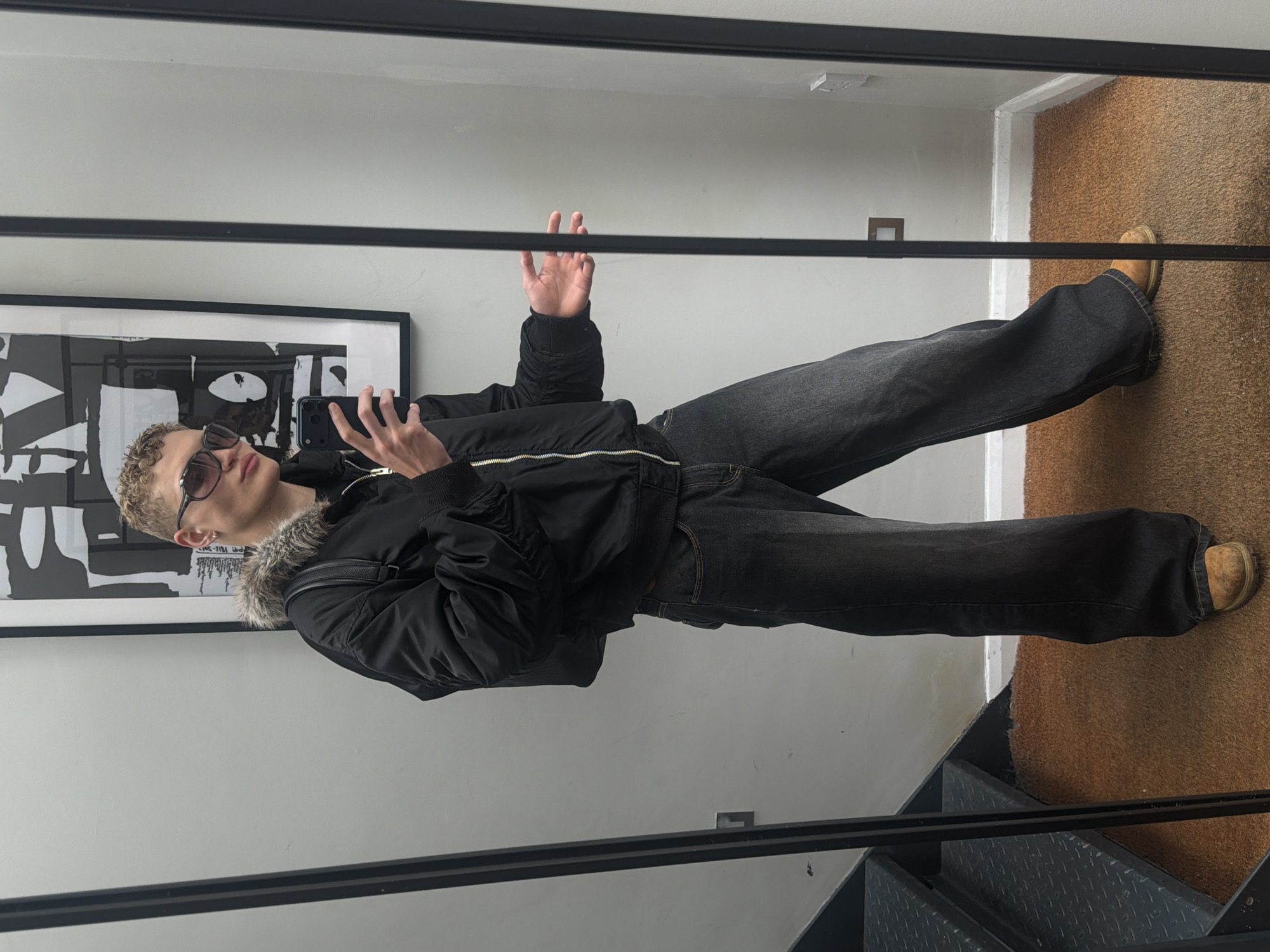 The image shows a person taking a mirror selfie. The person is wearing a black bomber jacket with a fur-lined hood, dark wash flared jeans, and tan shoes. They have blonde, curly hair and are wearing sunglasses. The person is holding a black smartphone in their right hand, which they are using to take the photo. In the background, there is a large framed black and white artwork on a light gray wall. The floor is covered with a brown carpet, and a metallic staircase is visible in the lower right corner. The overall lighting suggests an indoor setting, possibly during the daytime. The person's pose and the act of taking a selfie indicate a casual, everyday activity.