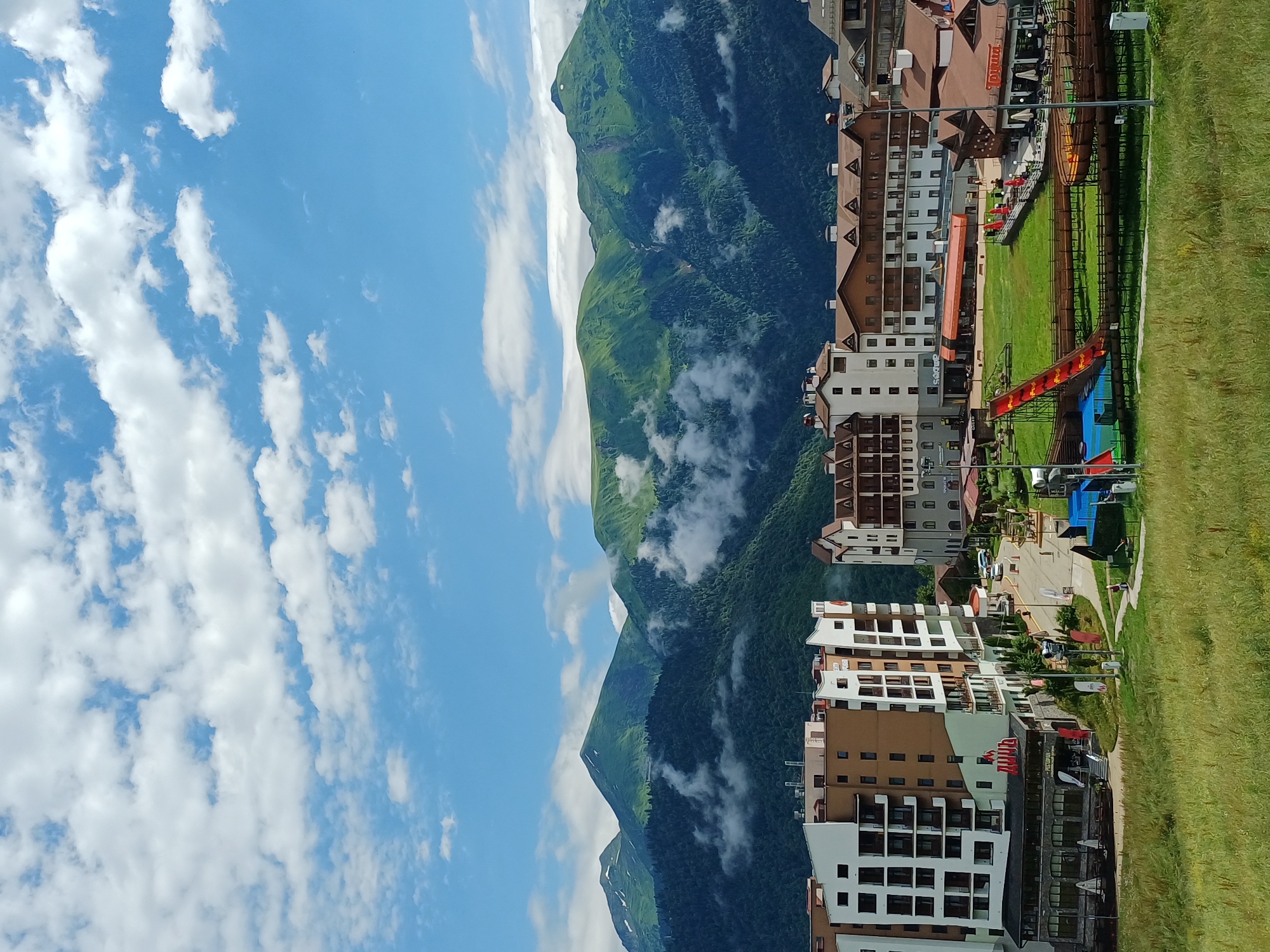 This image captures a panoramic view of the mountain resort town of Estosadok, Russia, under a partly cloudy sky. In the foreground, a grassy meadow slopes upwards, leading to a cluster of modern resort buildings. These buildings are multi-story structures with a mix of white, brown, and beige facades, featuring numerous balconies and pitched brown roofs. Some of the buildings have visible signage, including what appears to be a restaurant with outdoor seating.
Further into the scene, nestled between the resort buildings and the majestic mountains, a vibrant green lawn area is visible, populated with recreational facilities. A prominent red slide with a yellow track is a central feature, suggesting a playground or amusement area. Wooden pathways and fences create a network throughout this recreational space. In the background, a formidable mountain range dominates the horizon, their lush green slopes partially shrouded in wispy clouds. The peaks of these mountains are varied in height and shape, with some showing hints of snow. The overall scene depicts a well-developed tourist destination blending natural beauty with modern infrastructure.