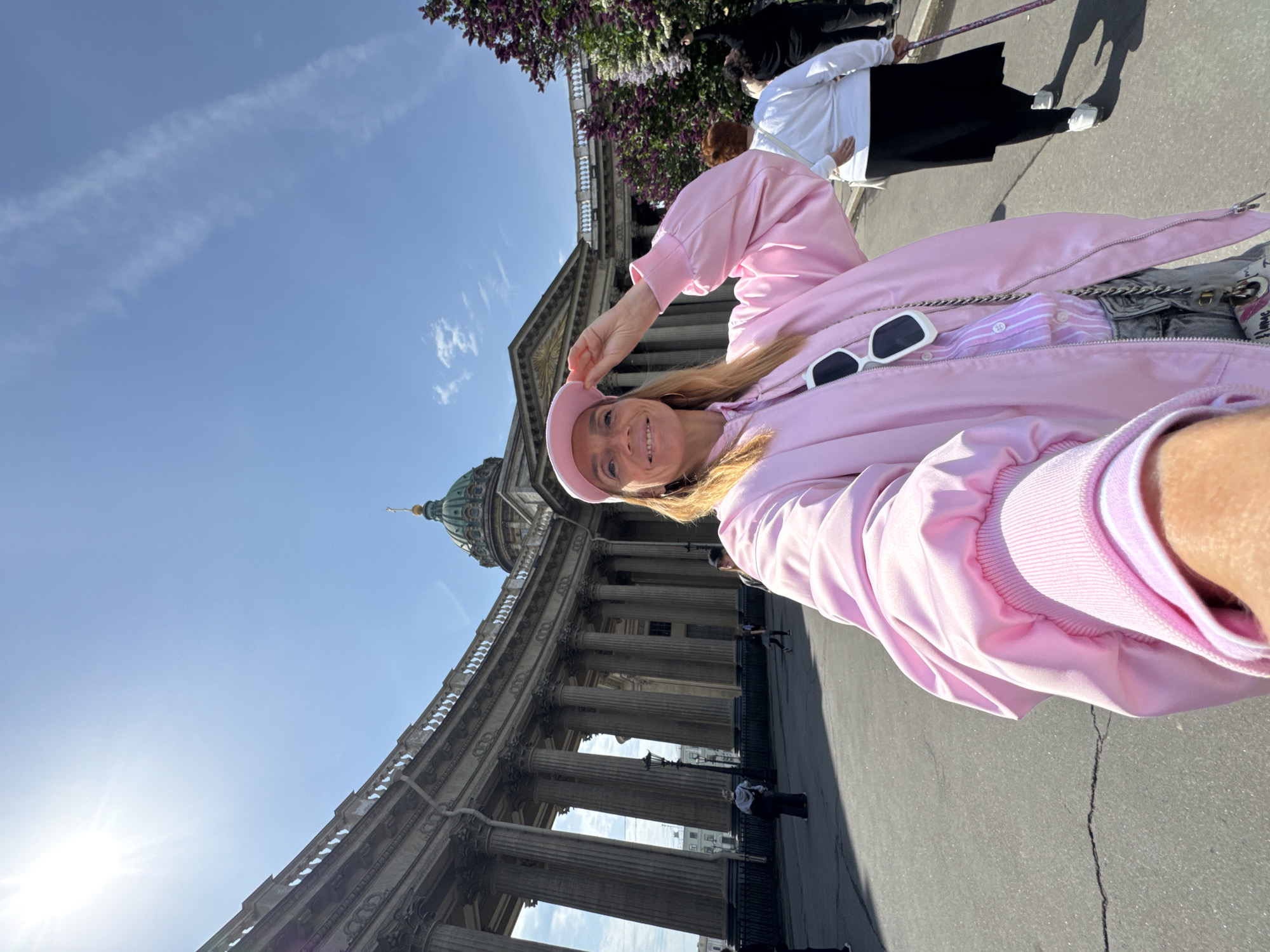 The image shows a woman in a pink outfit, including a beret, standing in front of Kazan Cathedral in Saint Petersburg, Russia. She is holding her beret and smiling. The cathedral's dome and colonnade are visible behind her. Other people are walking nearby. The sky is blue with light clouds. The sun is shining.