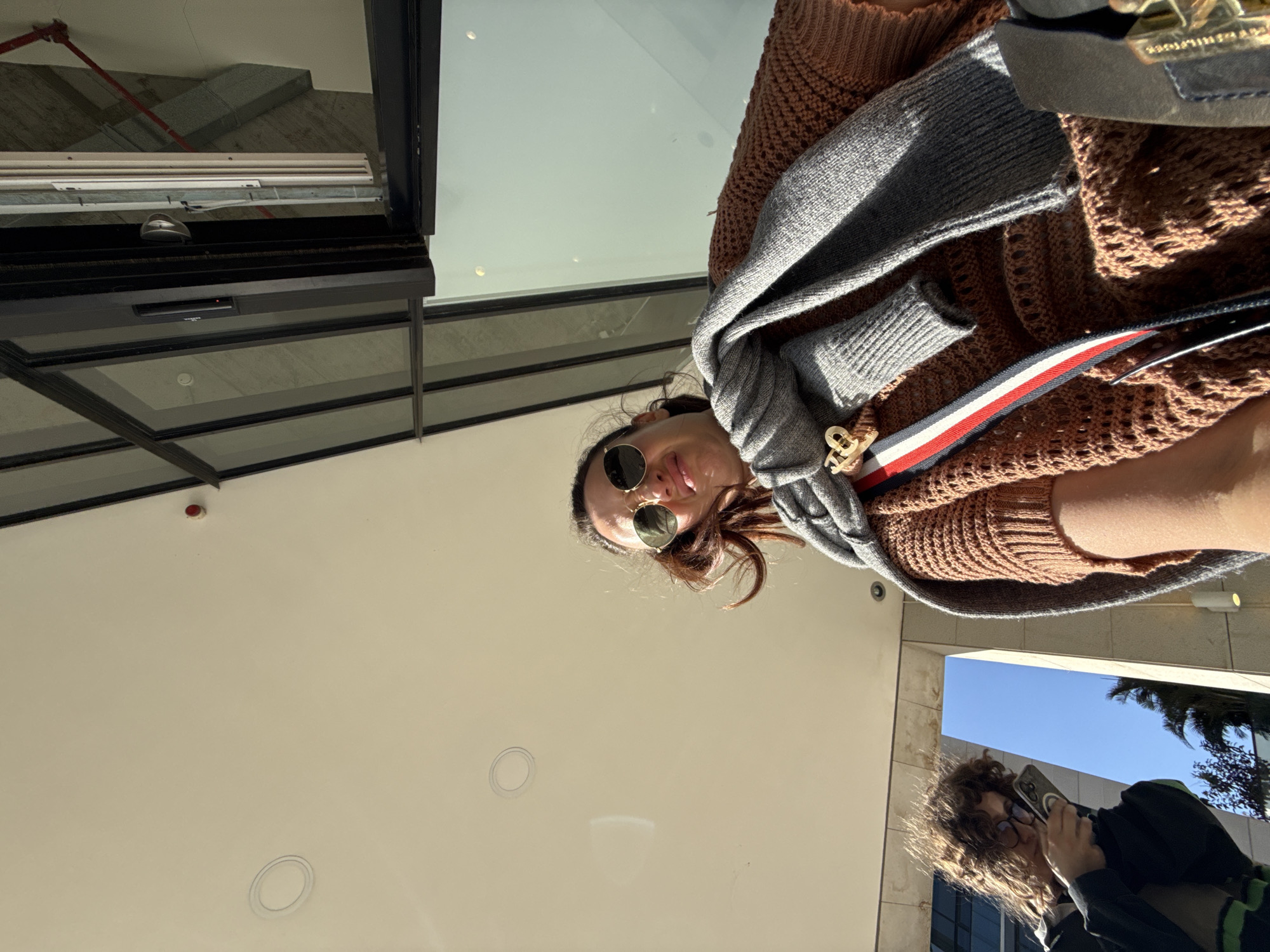 The image presents a low-angle perspective, likely taken with a camera or phone held pointing upwards.
**People:** Two individuals are visible. One is the primary subject, wearing sunglasses, a brown sweater, and a grey scarf. The second person is captured in the lower right corner, holding a smartphone and appearing to take a picture.
**Objects:** Sunglasses, a brown knit sweater, a grey scarf, a handbag, a smartphone, architectural ceiling elements including overhead lighting and a sprinkler system.
**Environment:** The setting appears to be an outdoor urban area, possibly Tel-Aviv based on architectural cues and common urban design elements. The ceiling and upper section of a building structure are visible, suggesting an open or semi-covered space. A portion of the sky and palm trees are visible in the lower-right corner, further supporting the location.
**Actions/Interactions:** The first person appears to be posing or looking at the camera, while the second person is actively taking a picture.