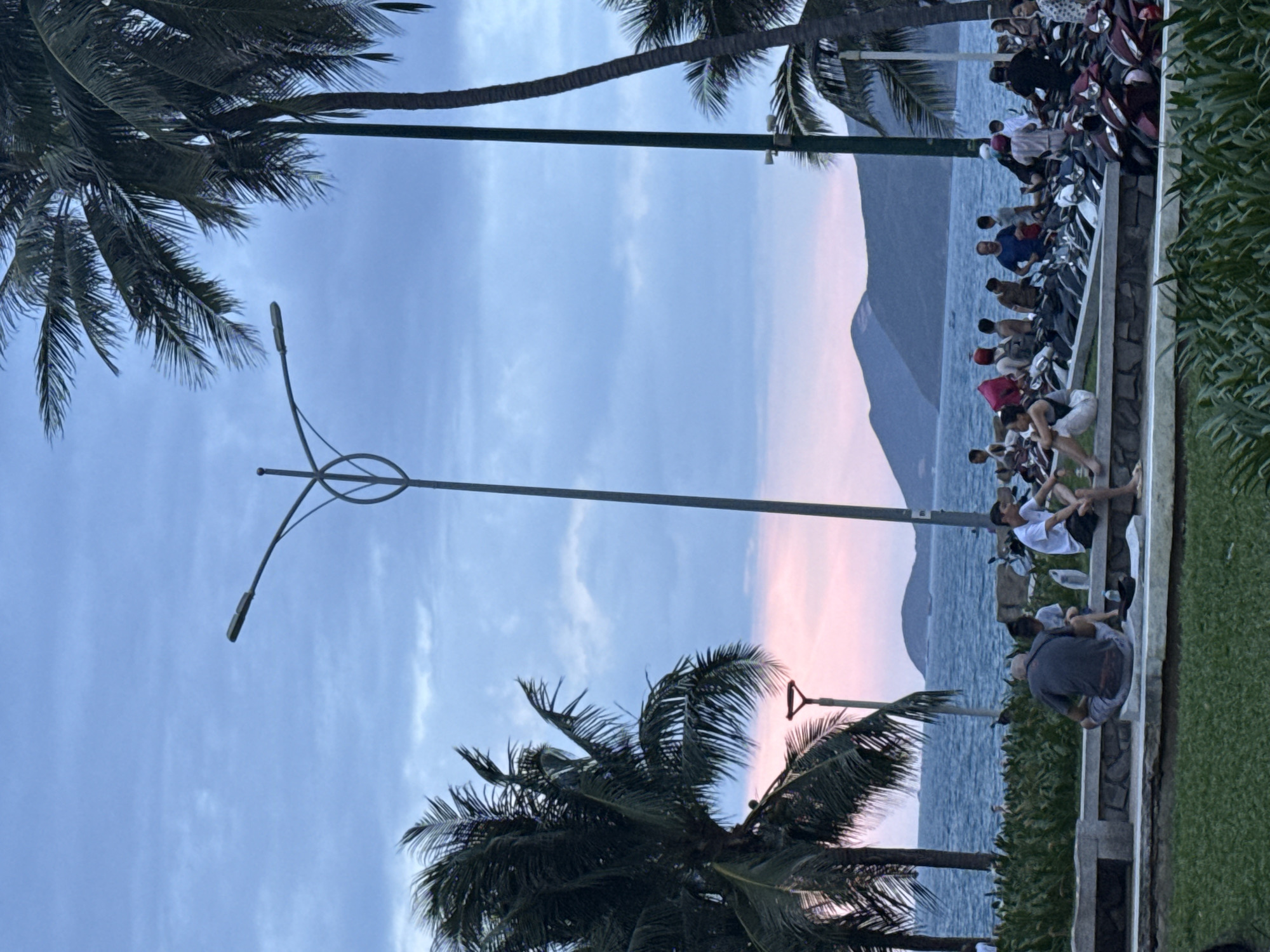 The image depicts a coastal scene in Nha Trang Ward, Vietnam. A group of people are sitting and interacting on a low wall along the beach, adjacent to parked motorbikes. The foreground includes palm trees and a street light. In the background, the ocean meets a mountain ridge, with a strip of pink sky indicating either sunrise or sunset.
