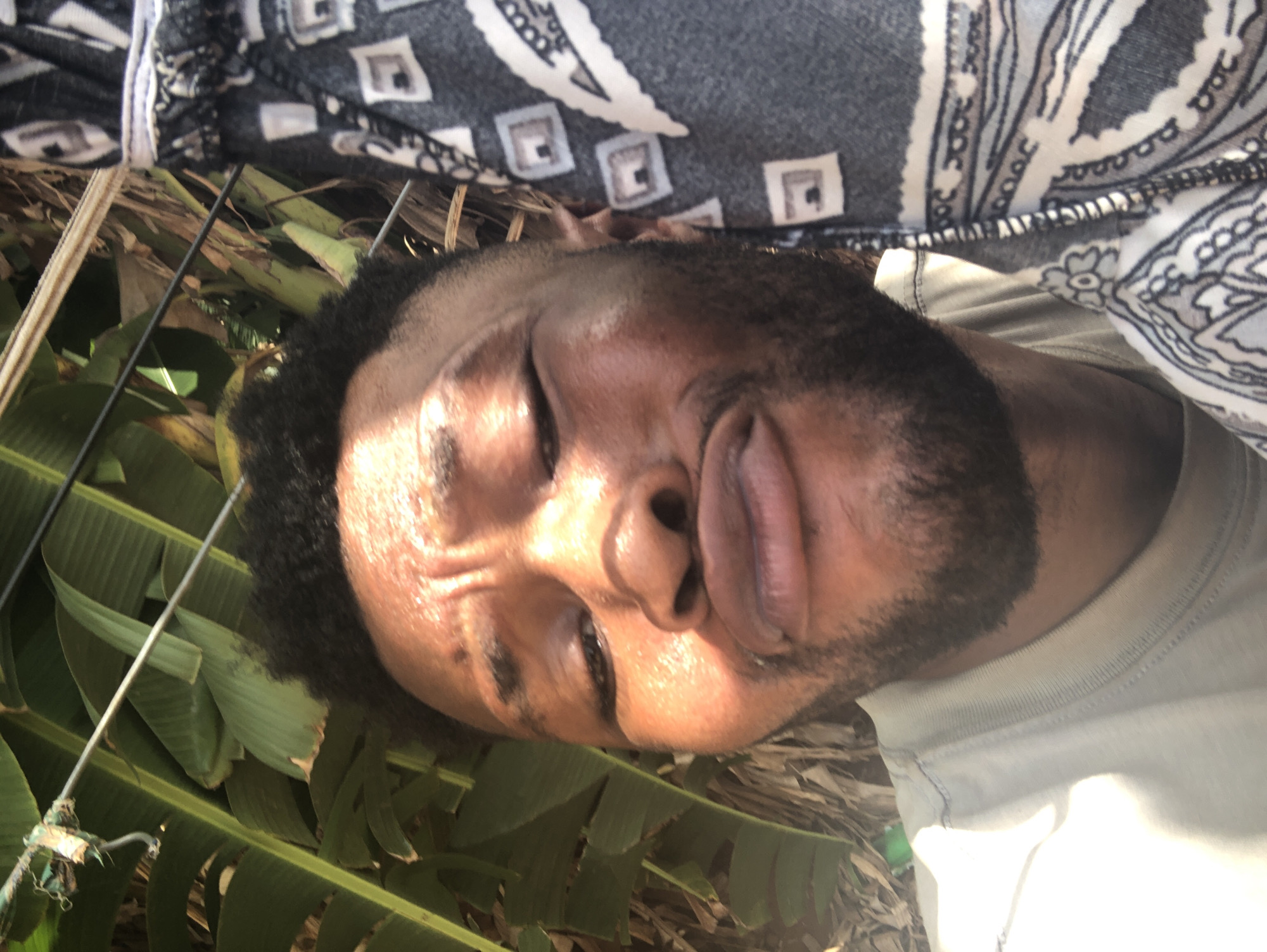 A close-up, slightly angled shot shows a man with dark hair and a beard lying down. His eyes are closed. The background consists of green banana leaves and what appears to be a fabric with a geometric pattern. The exact location and city are unknown.