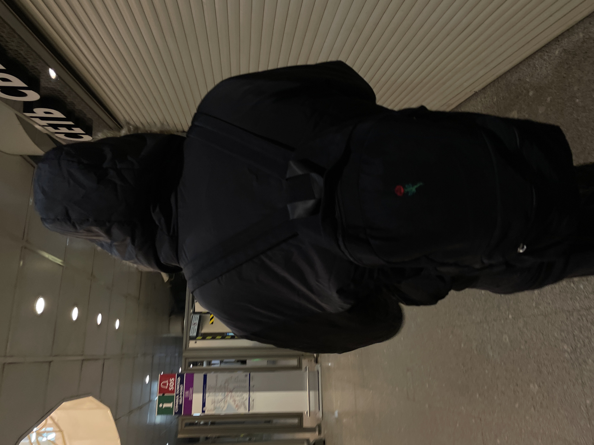 The image shows a person from behind, wearing a black hooded jacket and a black backpack. The hood of the jacket appears to have some fur or fleece lining. A distinctive red and green rose logo or patch is visible on the side pocket of the backpack. The person is walking or standing in what appears to be an indoor public space, likely a subway station or an underground passage, consistent with the provided location context of Saint Petersburg, Russia.
The ceiling features horizontal paneling and bright recessed lights. The walls are light-colored with similar paneling on the upper sections. The floor is a light grey, possibly concrete or tiled surface. On the left side of the frame, a black sign with white text, partially cropped, reads "СЕТЬ СВ" (SEТЬ SV). Further down a corridor, more public informational signage is visible, including a red square sign with "SOS" and a white square sign with a blue "i" for information. The overall lighting is artificial and somewhat dim, typical of an underground transit environment. The image is rotated 90 degrees clockwise.