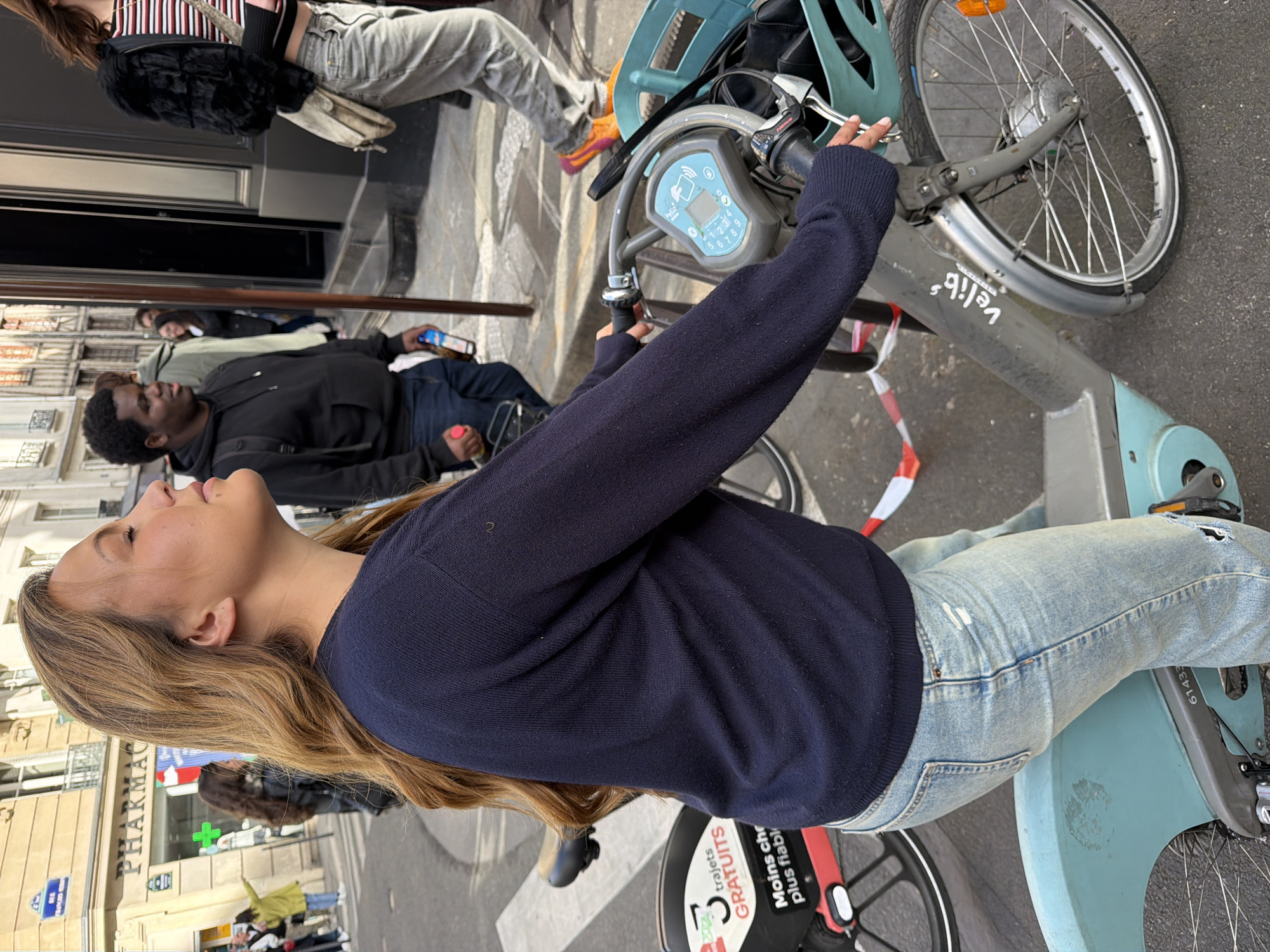 A young woman with long brown hair is sitting on a light-blue "Velib'" rental bicycle. She is wearing a dark blue sweater and light-blue jeans with a hole in the knee. Her head is tilted upward. The bicycle is parked on a street in front of a building with a "PHARMACIE" sign. A man is lying on the ground in the background, and other people are visible on the sidewalk. A sign on the bicycle reads "trajets GRATUITS" in red letters.