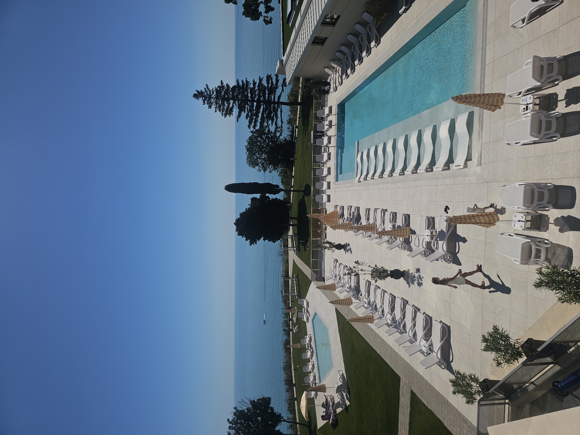 The image shows an aerial view of a resort swimming pool area on a clear, sunny day. The large rectangular pool is filled with blue water and has a shallow section with built-in lounge chairs. The surrounding patio is tiled and lined with white lounge chairs and yellow and white striped umbrellas. A woman in a white dress is walking across the patio. In the background, a large expanse of the blue ocean stretches to the horizon. Trees and green grass are visible along the coastline.