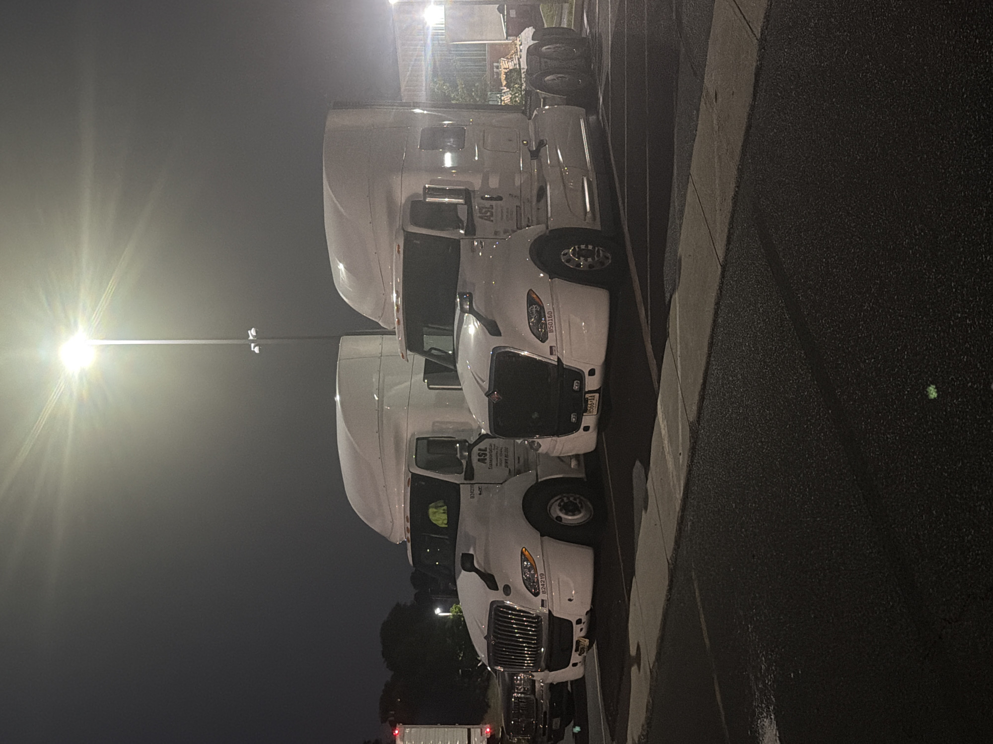Two white semi-trailer trucks are parked in a lot at night, illuminated by a bright overhead light. The truck on the left is a 2020 International LT, with the logo "ASL Transportation" visible on its side. The truck on the right is a newer model, also white with the "ASL Transportation" logo. Both trucks are clean and appear to be in good condition. The parking lot is paved and appears to be empty except for the trucks. In the background, there are some trees and buildings, as well as other vehicles. The sky is dark and clear, with no visible stars. The overall impression is one of a quiet, industrial area at rest. The image is undated and no specific location is provided beyond "New Britain, United States of America".