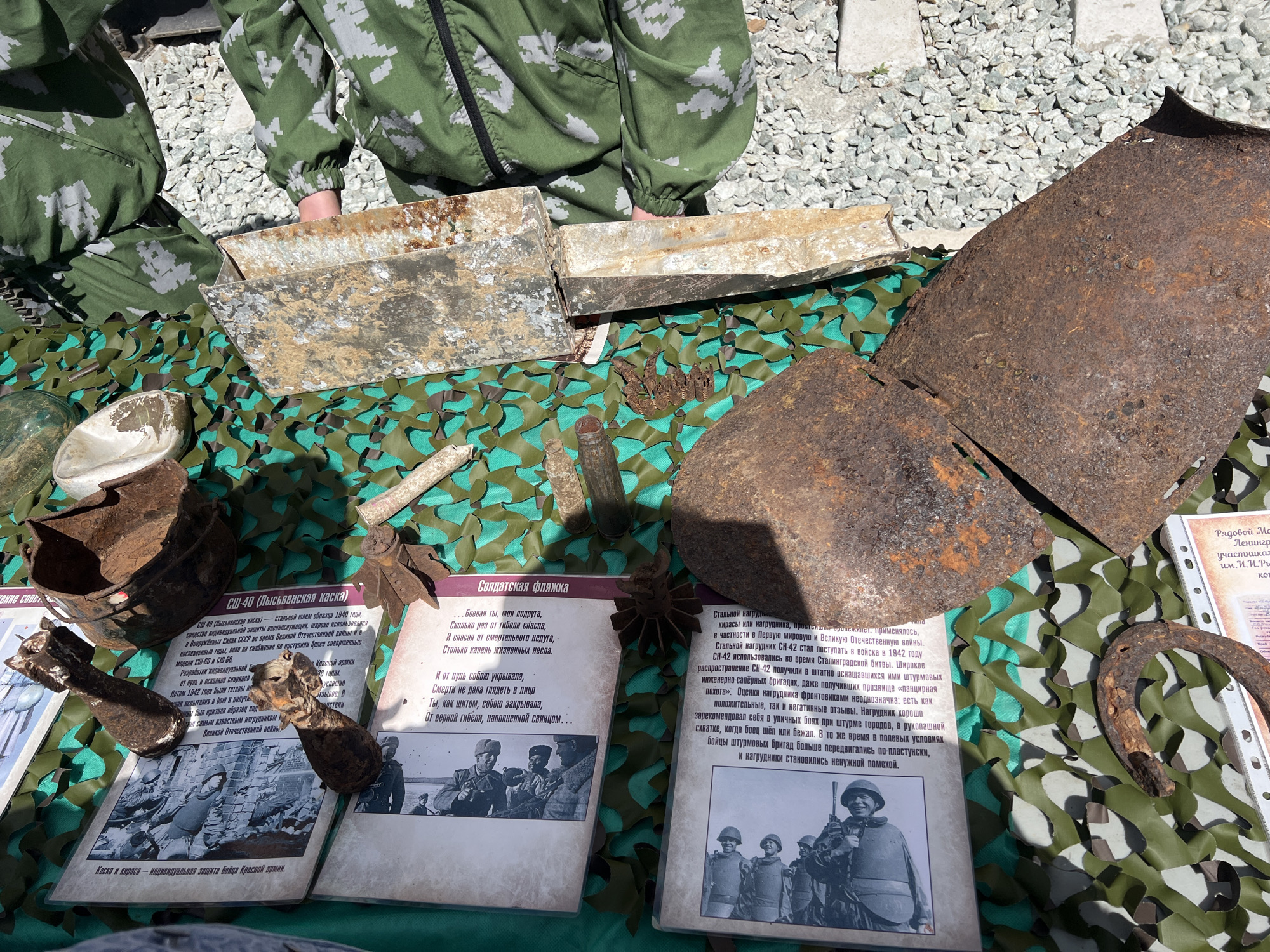 The image shows a display of historical military artifacts, likely from World War II, set up outdoors, possibly in Ufa, Russia. The artifacts include rusty metal helmets, shell fragments, a Soviet flask, and ammunition. There are information cards written in Russian describing the objects, such as the SSh-40 helmet and the "Soldier's Flask". A person in a green and gray camouflage suit is partially visible at the top of the image. The objects are displayed on a camouflage net placed over a table. A horseshoe is also included in the display.