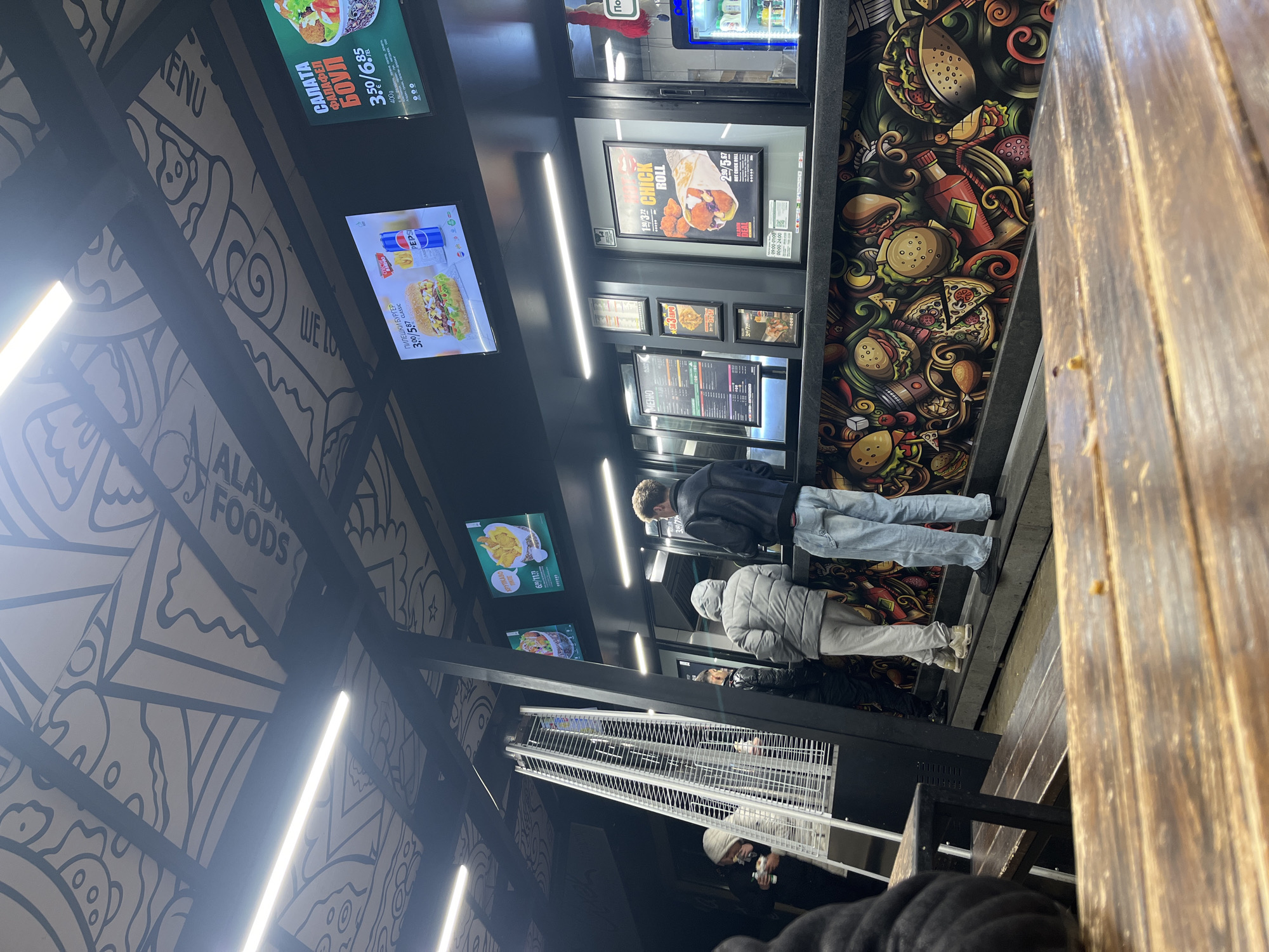 The scene appears to be the outdoor area of a fast-food establishment named "ALADIN FOODS" in an unknown city. People are queuing to place orders, indicated by the menu boards and digital displays overhead showing food items like "Салата Фалафел" (Salad Falafel) for 3,50/6.85, a chicken burger for 3,00/587, and a chicken roll for 2,90/5-67. The decoration includes a patterned wall with food-related doodles. There's also a wooden structure, possibly part of the seating or building facade. A metal cage-like structure and outdoor heaters suggest provisions for customers even during colder weather.