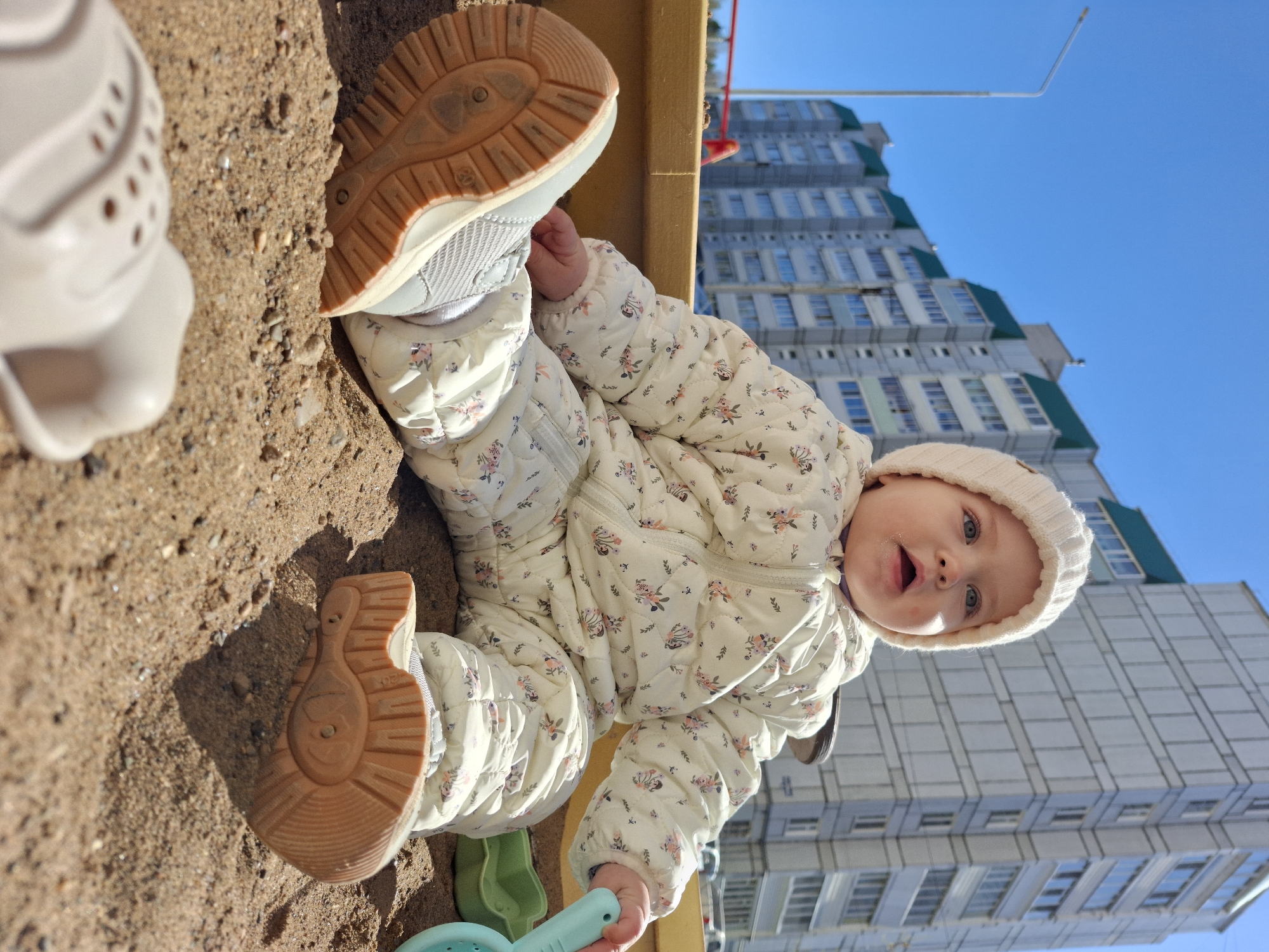 A baby in a white floral snowsuit and knitted hat is sitting in a sandbox, holding a blue and green plastic shovel. The baby is looking up with an open mouth. In the background, there is a tall apartment building with light gray walls and green roofs, under a bright blue sky. The photo was taken in Chaikovsky, Russia.