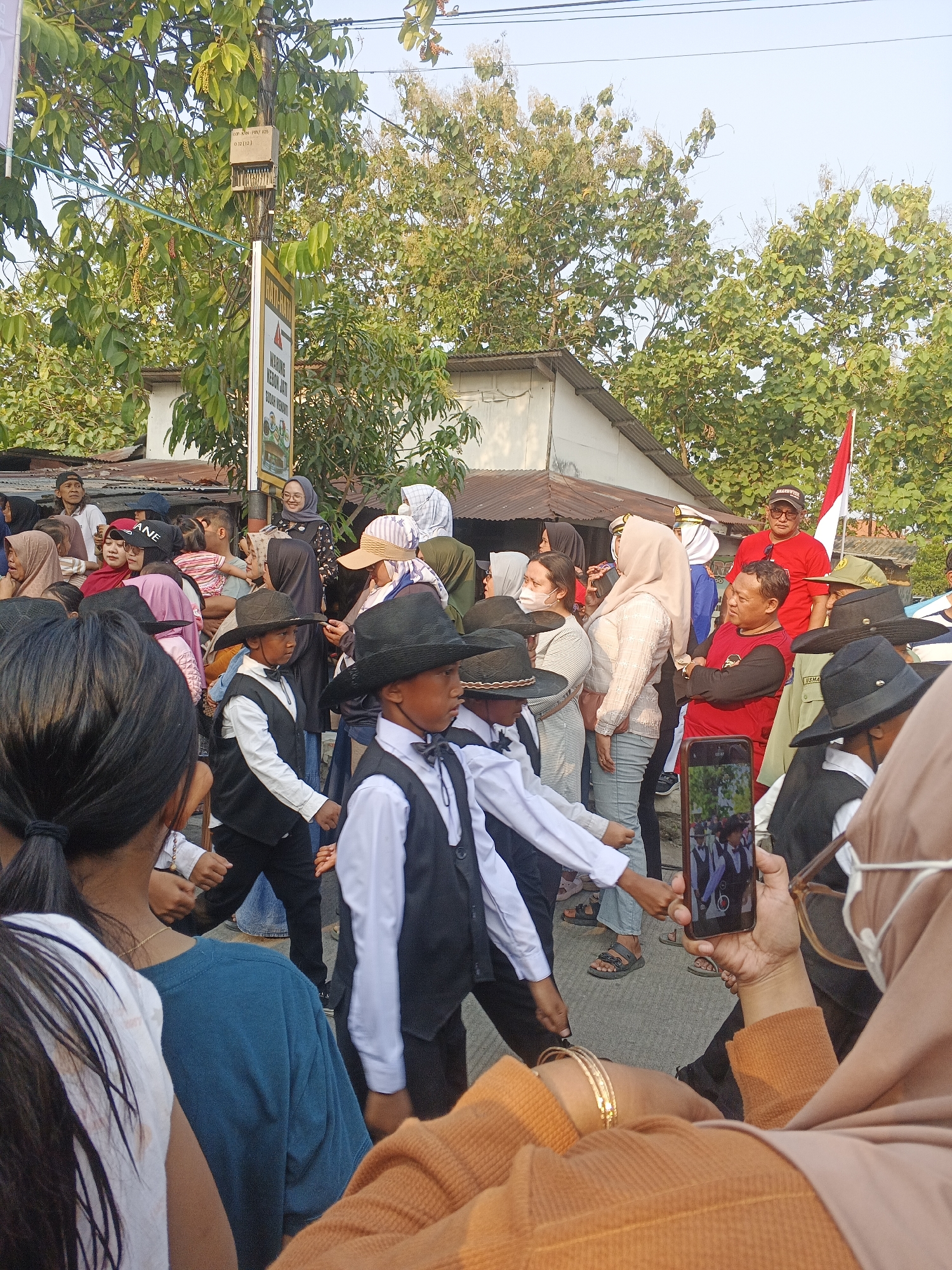 The image captures a street scene in Bogem Pinggir, Indonesia, during an outdoor event. A crowd of people, dressed in a variety of casual attire and headscarves, are gathered, some appearing to watch a procession or performance. Several young boys in the foreground are dressed formally in white shirts, black vests, and black cowboy hats, suggesting they are part of an organized group or parade. One of the boys is looking to the side, while another is facing forward. In the background, buildings with corrugated metal roofs are visible, alongside trees and utility poles with electrical wires. A red and white Indonesian flag is planted on a pole. A woman in the foreground is holding a smartphone, displaying a photo of the young boys in their formal attire, possibly capturing or reviewing the event. The lighting suggests it is daytime, with the sun casting shadows.