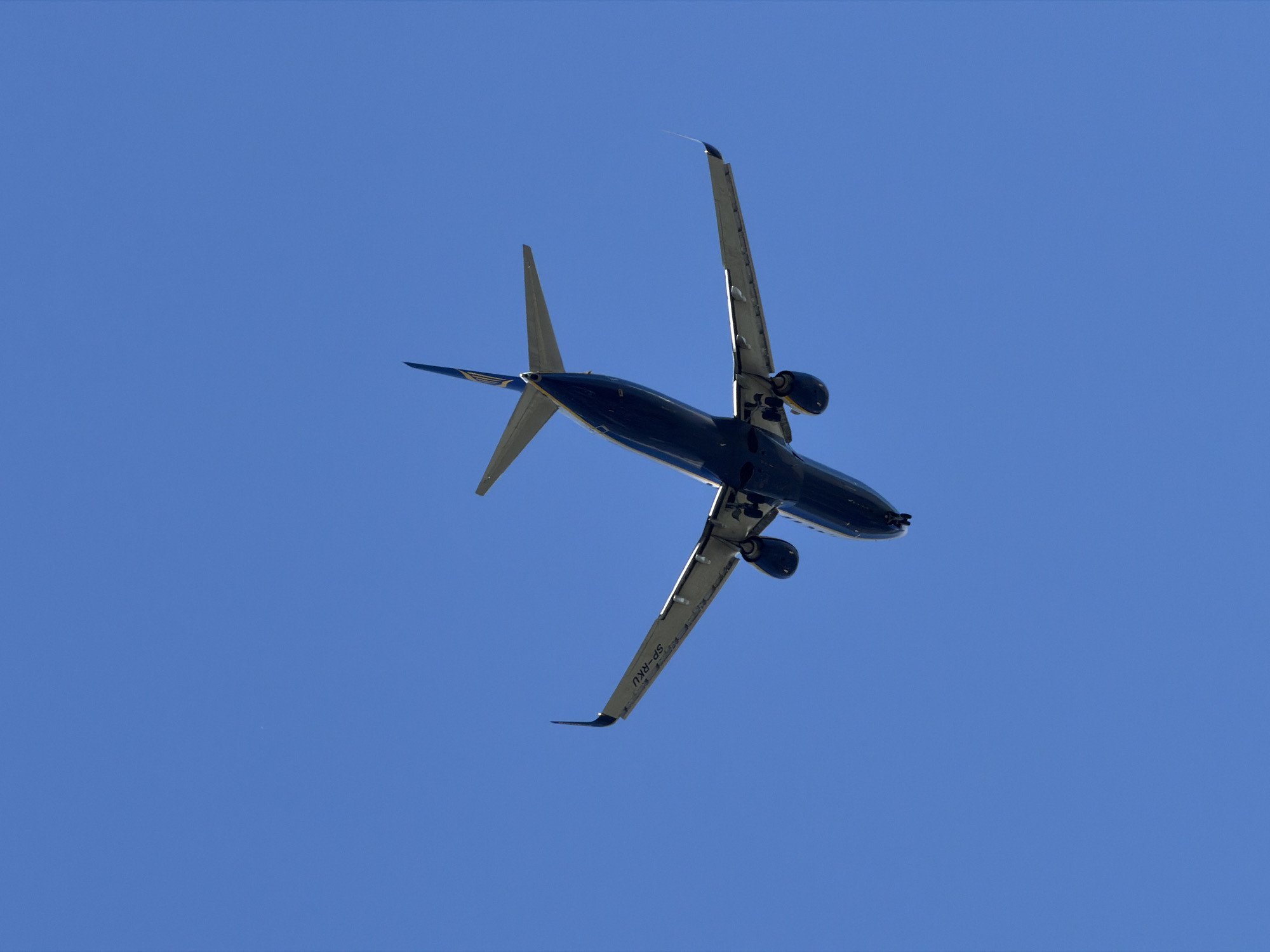 An underside view of a dark blue airplane with yellow accents is captured against a clear, bright blue sky. The aircraft is in flight, with its wings extended and engines visible. The registration number "SP-RKU" is visible on the left wing. The plane appears to be approaching or departing from an airport. The image does not provide specific visual cues to determine its exact location within the city of Burgas, Bulgaria, beyond it being in the sky above the city.