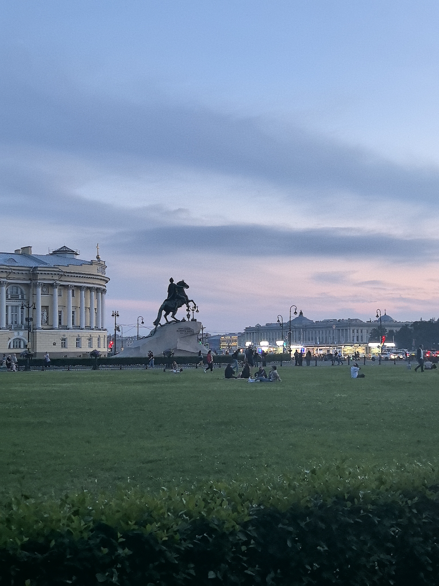 The image depicts the Bronze Horseman, a monument to Peter the Great, in Senate Square in Saint Petersburg, Russia. The equestrian statue is prominently displayed on a granite pedestal against a dusky sky with wisps of pink and blue clouds. In the background, the pale yellow facade of a building with classical columns stands to the left, and further in the distance, a long, neoclassical building with illuminated windows and domes is visible. A wide expanse of green grass, where several people are seated and standing, occupies the foreground and middle ground. A low hedge runs along the bottom of the image. The overall scene suggests a public gathering in a city square during twilight.
