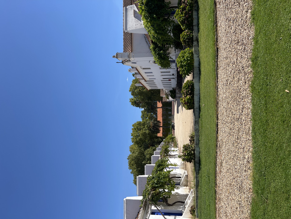 The image displays an architectural complex in Coruche, Portugal, under clear blue skies and bright daylight. The scene is presented from a low-angle perspective, looking into a central, unpaved courtyard.
The foreground consists of three distinct horizontal layers: a strip of manicured green grass at the bottom, followed by a wider band of light-colored gravel, and then another strip of green grass, slightly elevated and bordered by a low concrete barrier. On this elevated grass, a series of rounded, leafy green bushes are arranged in front of the buildings.
The midground is dominated by a large, open courtyard made of light brown dirt or gravel. Flanking this courtyard are two sets of white-painted buildings with terracotta-tiled roofs. On the left, a row of single-story structures features blue-painted arched doorways and windows. Green, leafy vines are trained on trellises or pergolas along the front of these buildings. On the right, a taller, multi-story white building with a terracotta roof presents a more elaborate facade, including a gabled section topped with a small cross-like ornament. Steps lead up to an entrance on this building, and green foliage also adorns its front.
In the background of the courtyard, further buildings with reddish-brown tiled roofs are visible, partially obscured by dense green trees, including a prominent palm tree. A dark-colored sedan is parked near the far-left building at the rear of the courtyard. No individuals are present in the scene.