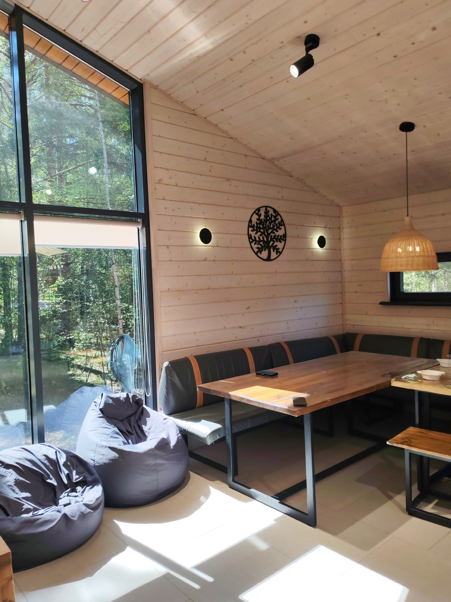 The interior of a room with wood paneled walls and ceiling features a corner seating arrangement consisting of a gray bench with orange accents against the wall and a wooden table with black metal legs. To the left, large windows provide a view of a lush green forest. Two gray beanbag chairs sit on the floor near the window. On the wall, there is a decorative metal tree and two sconce lights. A woven pendant lamp hangs above the table. The room is well-lit, with natural light streaming through the windows and artificial light from the fixtures. It is not possible to confirm the location in the city of Izhevsk, Russia.