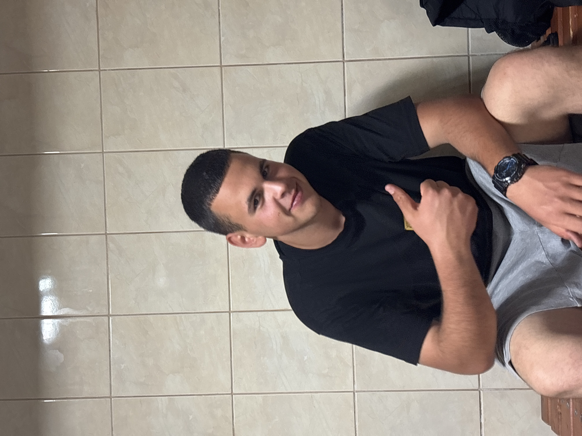 A young adult male is seen indoors, likely in a locker room, due to the tiled wall behind him. He has short, dark hair and wears a black t-shirt, grey shorts, and a large, dark-colored watch on his left wrist. He is lying down or sitting on something out of view. The subject is looking towards the camera and giving a thumbs-up gesture. No specific details are apparent to confirm the location within the city of Zlatograd, Bulgaria.